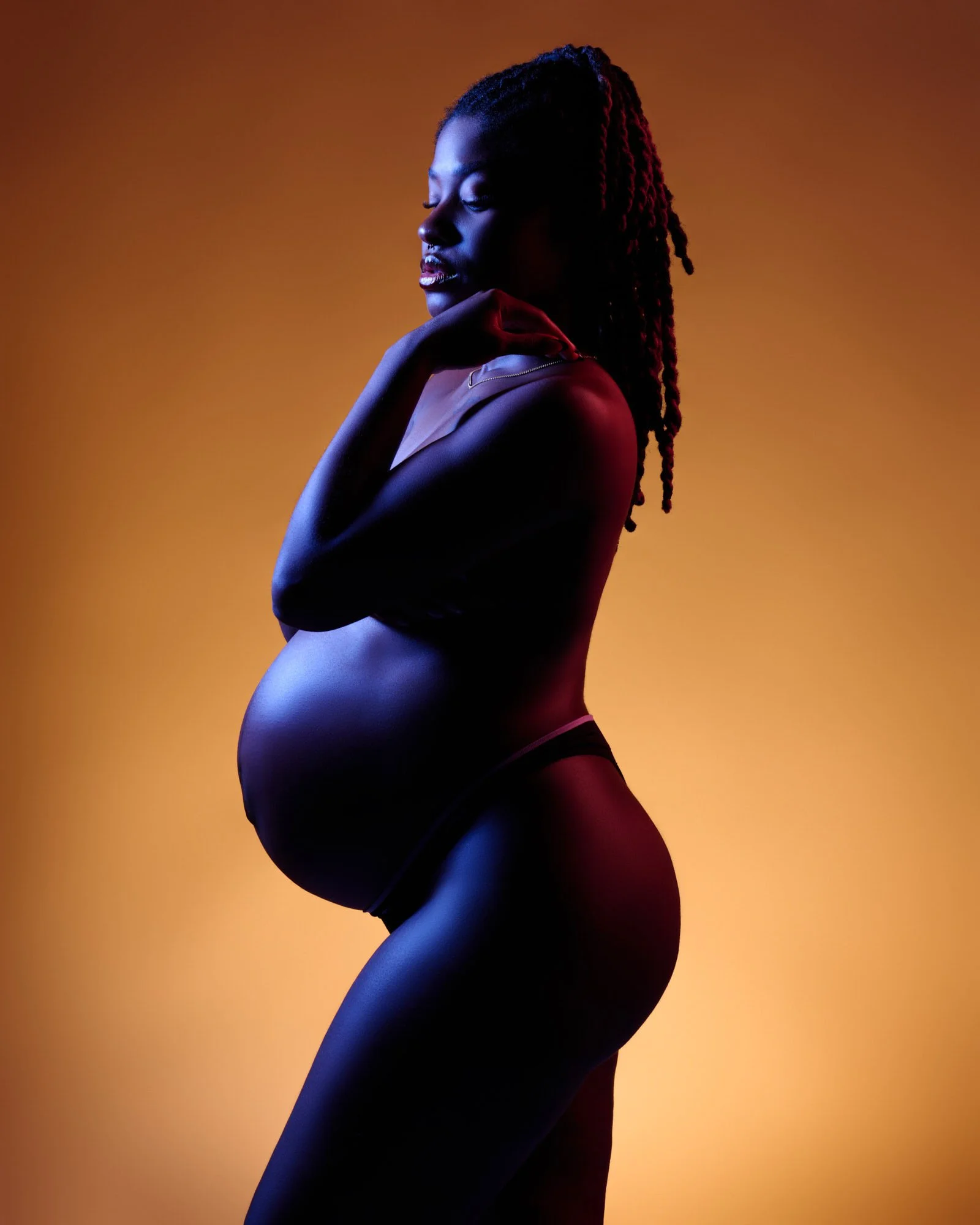Pregnant woman lit with orange and violet gels, hand at her chin, an edgy studio maternity portrait.