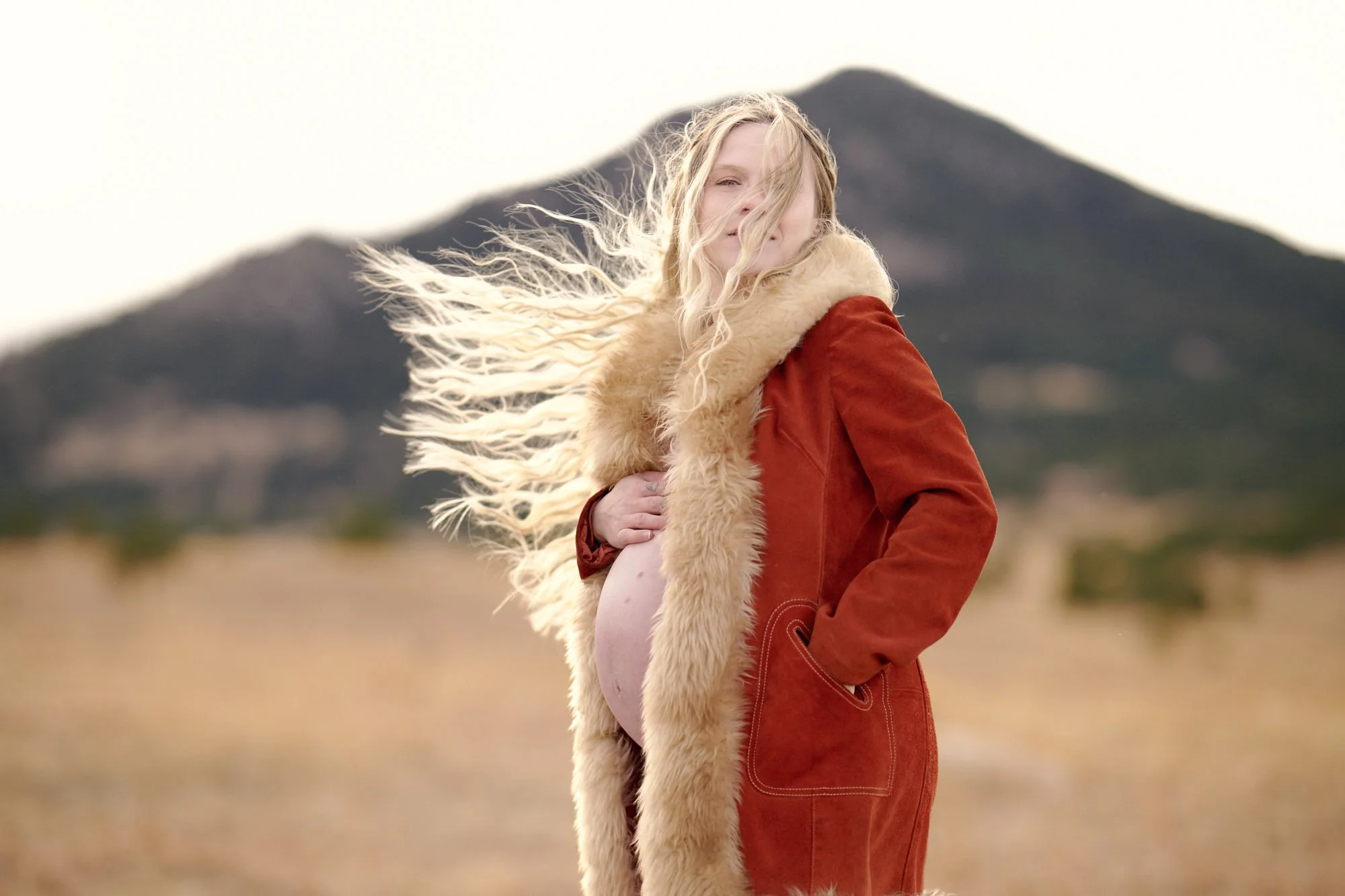 Rust suede coat with shearling trim, hair wild in the wind below a dark peak.