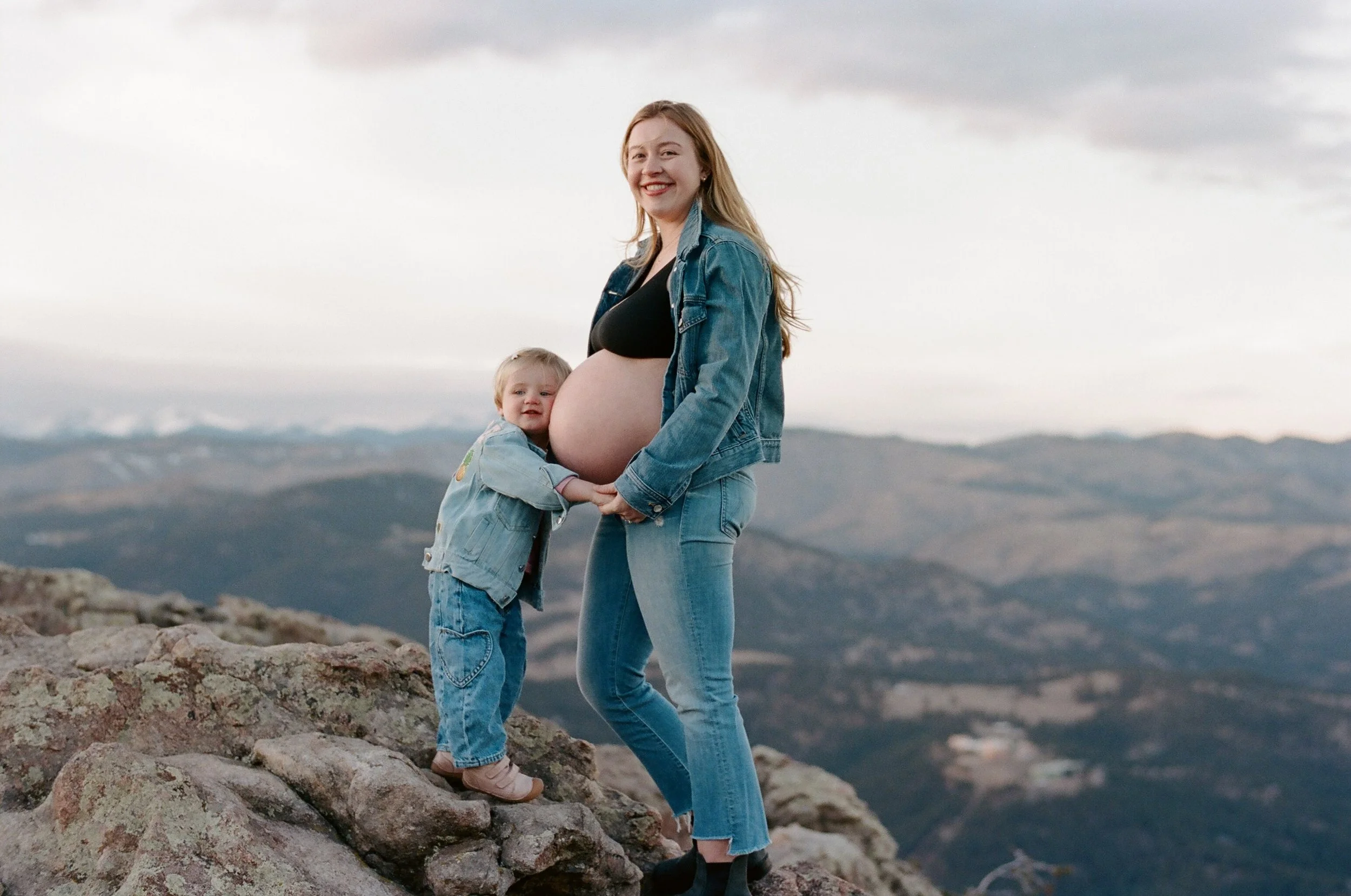 Smiling pregnant woman in denim holding hands with toddler on rocky mountain overlook