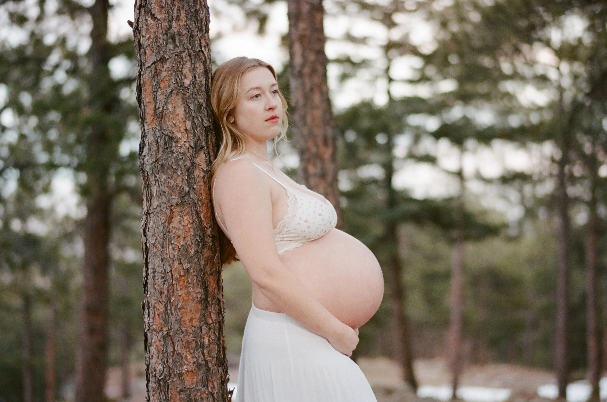 Leaning on a ponderosa in a white lace top and skirt, bare bump, winter forest on color film.