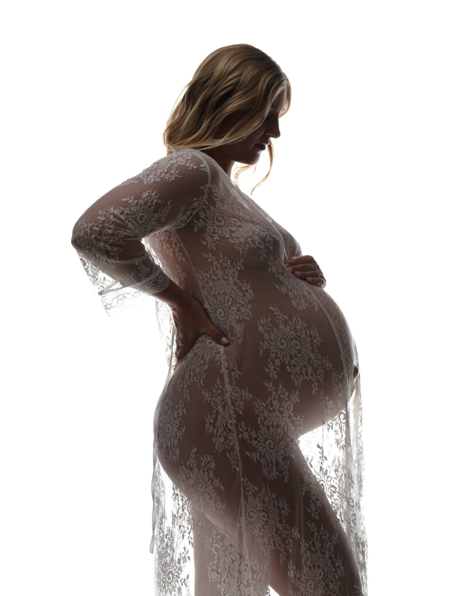 Sheer lace gown from behind, hands lifted to her hair in soft studio light.