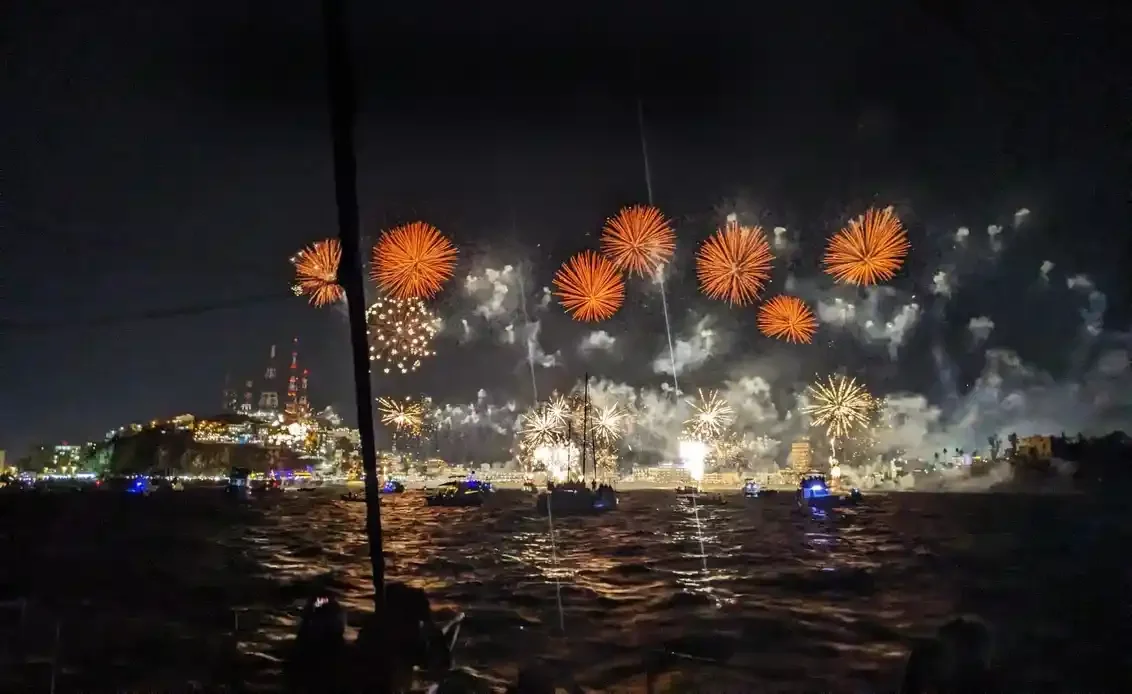Orange and white fireworks burst over the Pacific during the Combate Naval at Carnaval de Mazatlán, viewed from a sailboat with the city's skyline lit along the shoreline.
