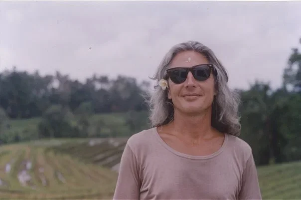 Randy in 1993 standing in front of Balinese rice fields, long grey hair loose, sunglasses on, a frangipani flower tucked behind one ear.