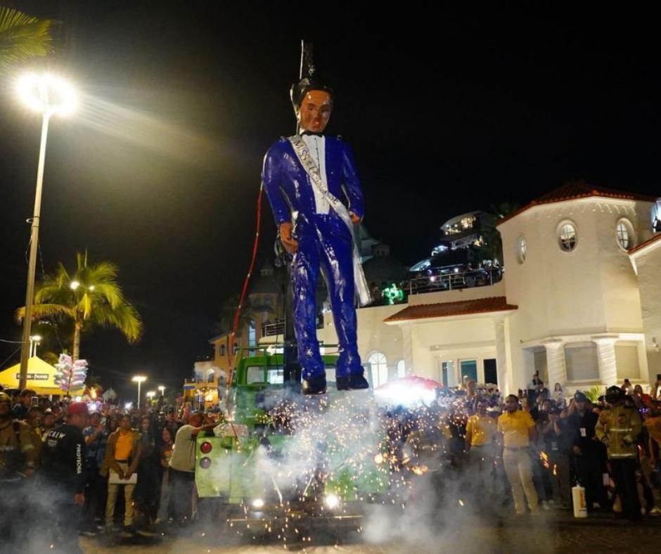 A tall blue-suited monigote effigy burning at night along the Mazatlán Malecón as fireworks scatter sparks around the crowd.