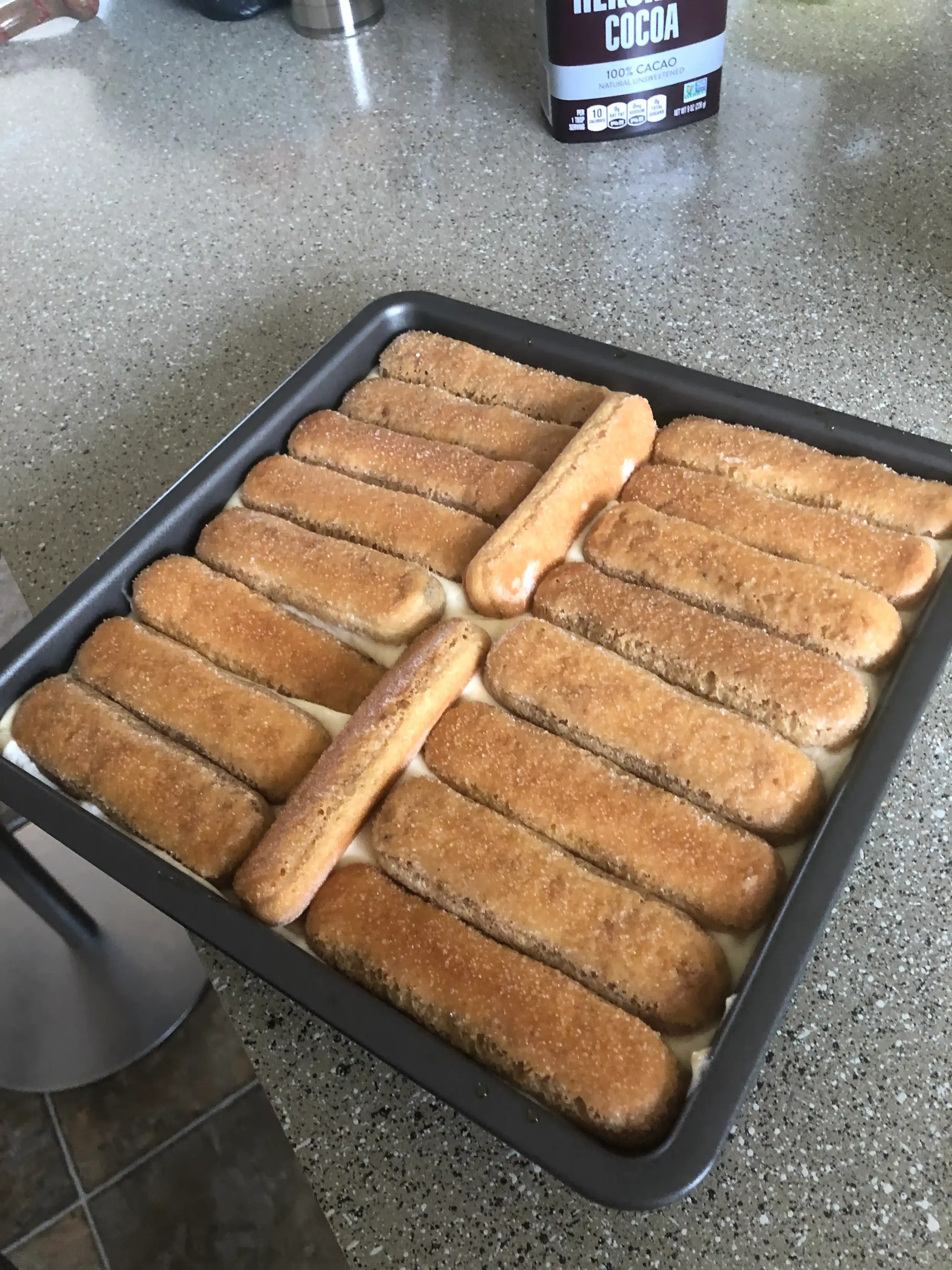 Savoiardi ladyfinger cookies arranged in a single layer in a square baking pan, ready for the second pour of cream.