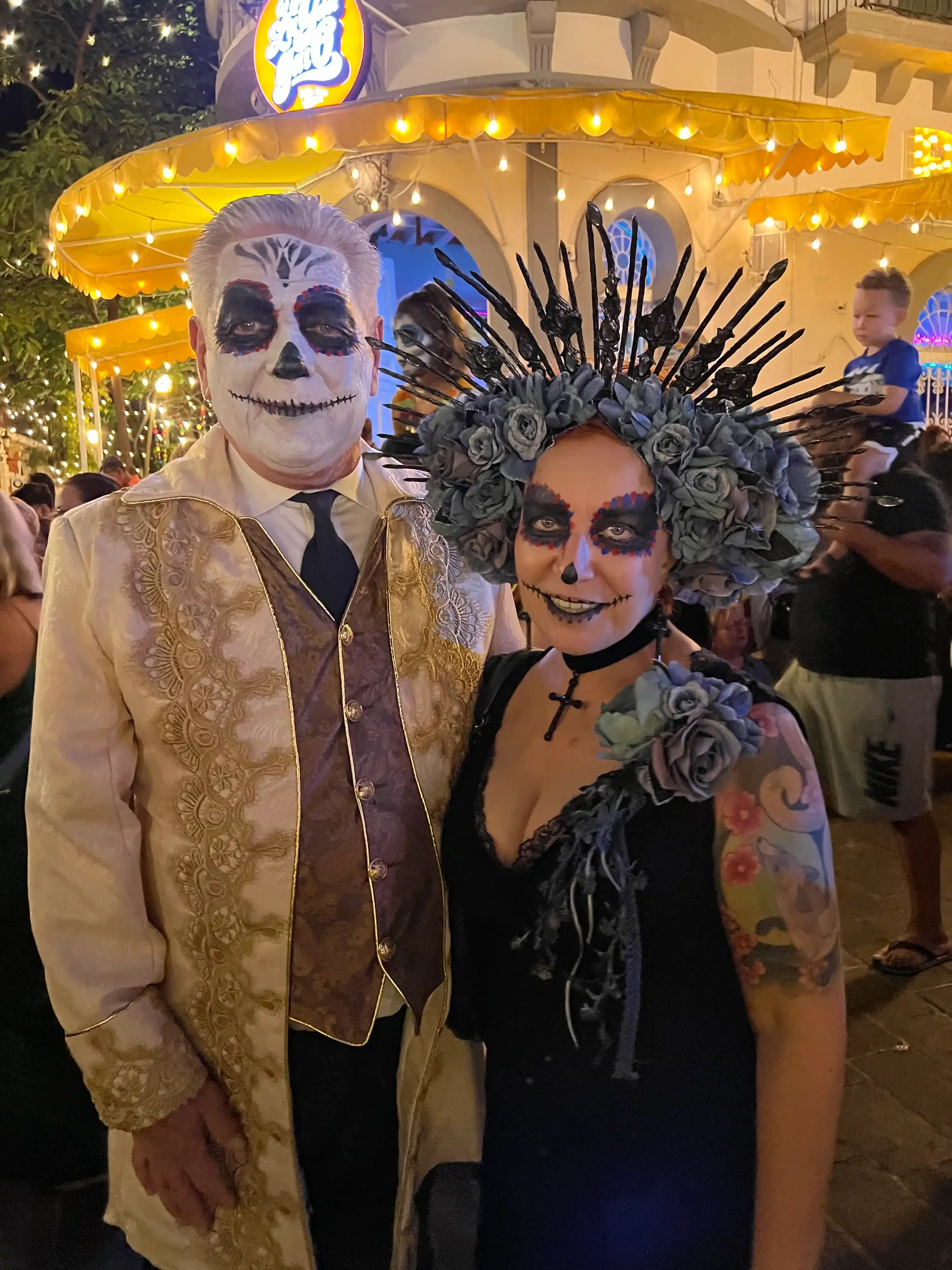 Inga and Randy in full sugar-skull face paint and Day of the Dead costume at the Mazatlán festival, the lit gazebo of Plaza Machado behind them.