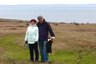Inga and her husband standing together in a wide open field of pale grass under an overcast sky, both in winter coats, on one of the many walks Rudy has joined over the years.