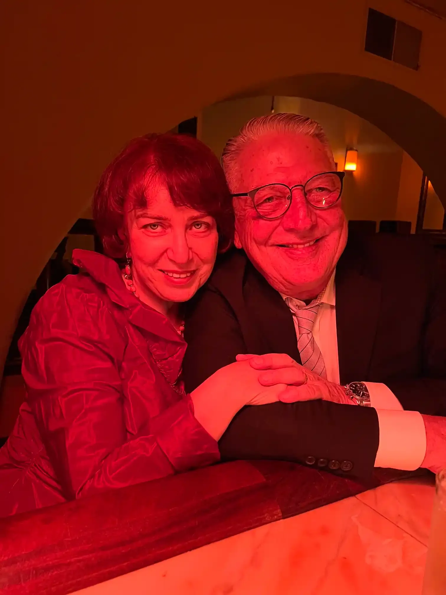 Inga and Randy at a candlelit table inside Il Bistro restaurant in Seattle, holding hands, photographed under warm red light.