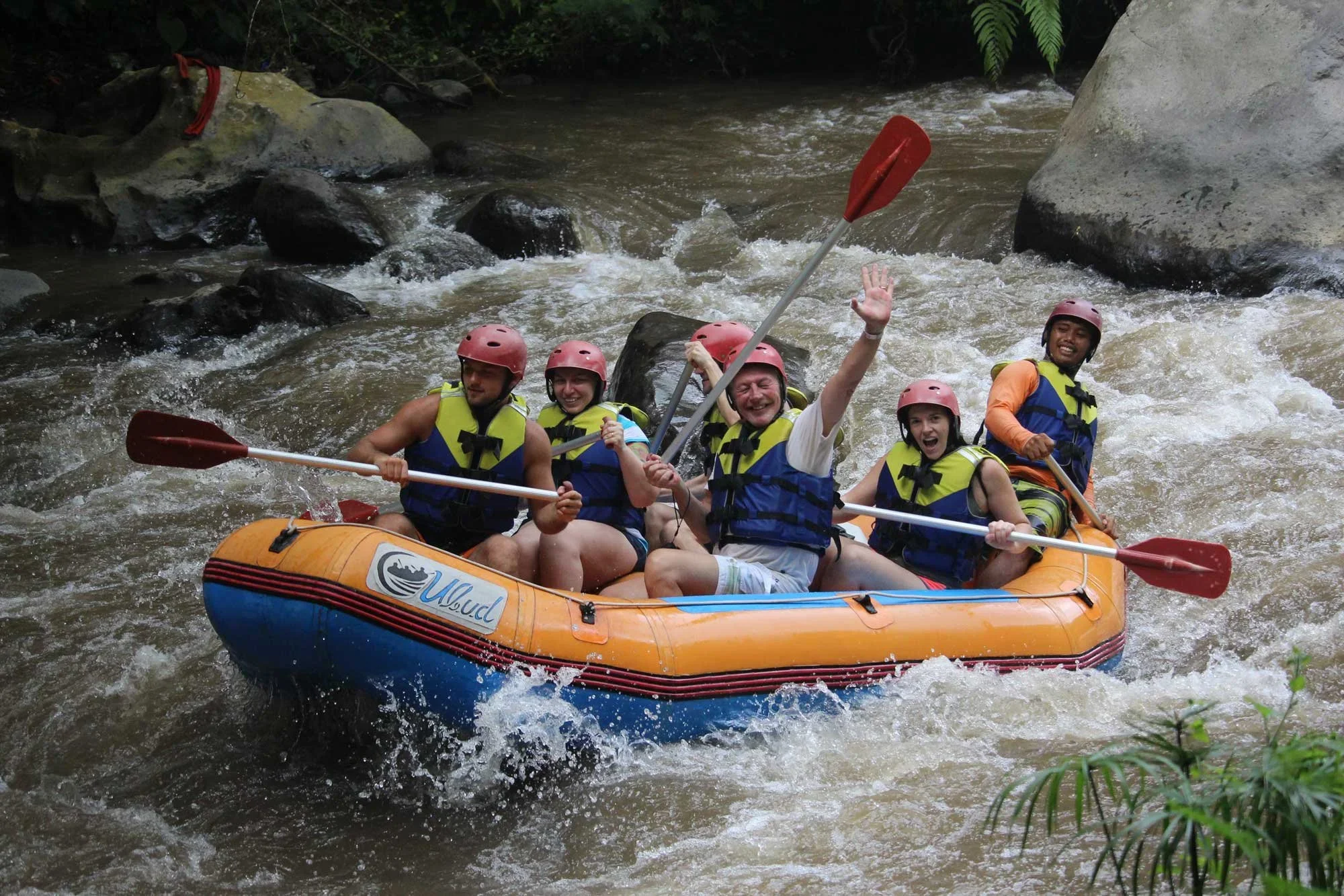 Five rafters in helmets and life vests grinning and raising paddles as their orange raft hits white water on a jungle river in Bali.