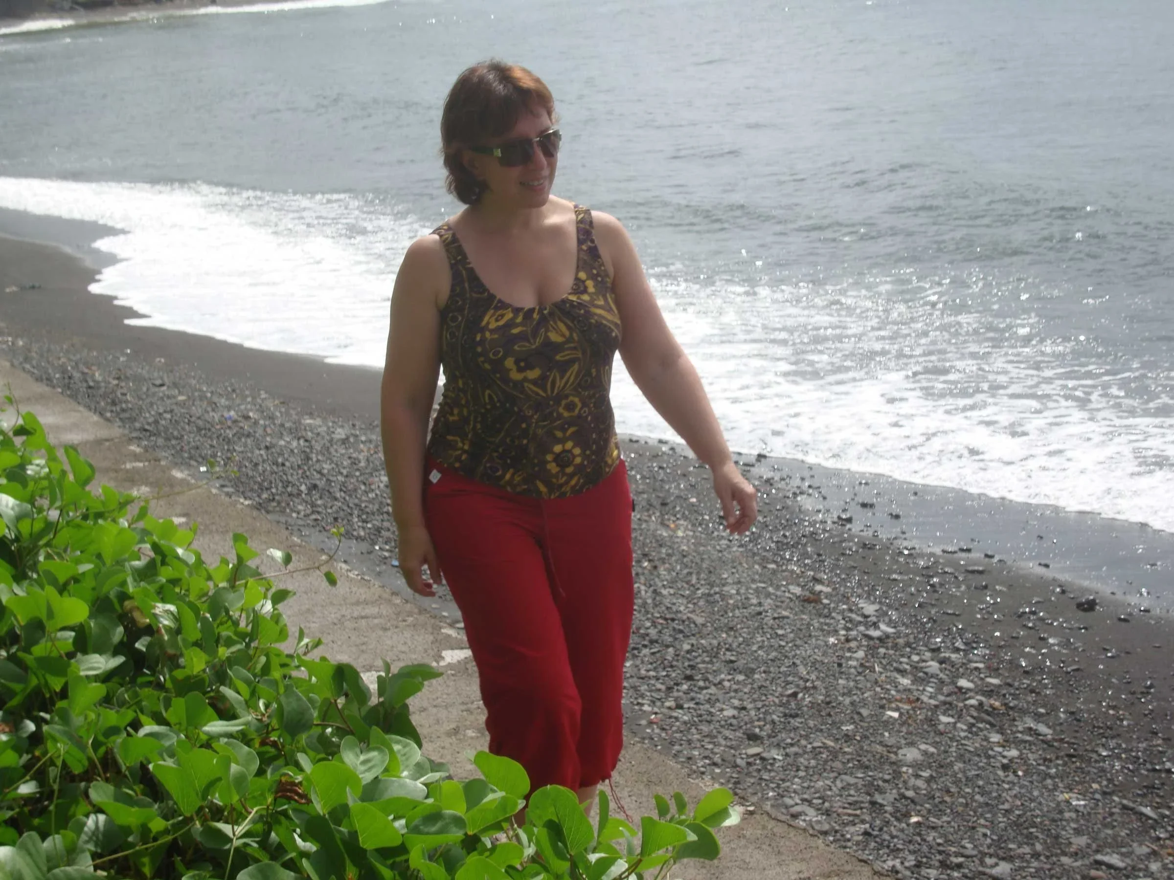 Inga in a patterned top and red trousers walking along Lovina's dark sand beach in north Bali as a wave breaks behind her.