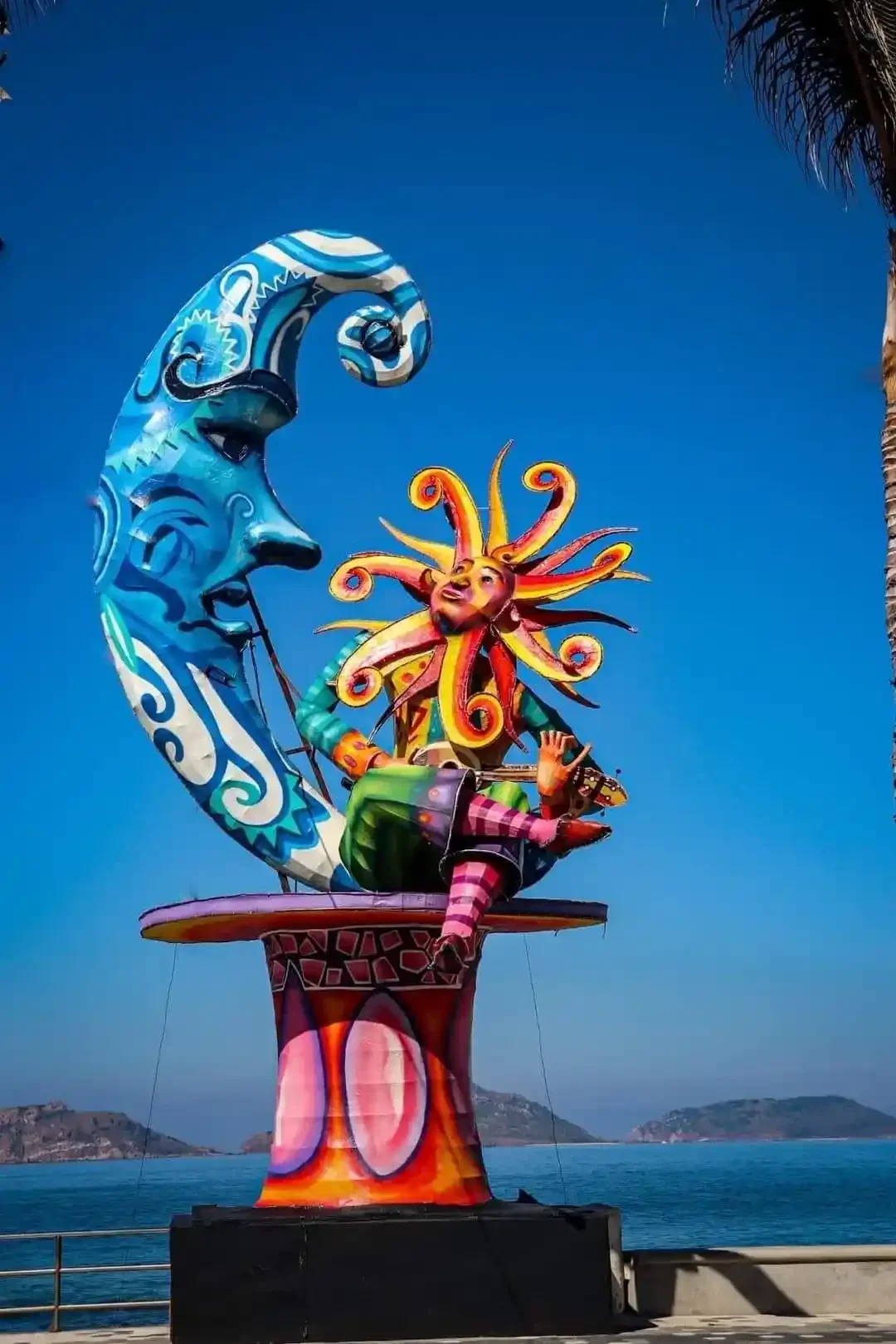 A monigote on the Malecón depicting a colorful sun figure with a wild orange and yellow mane lounging against a tall blue crescent moon, set against the Pacific Ocean and distant hills.