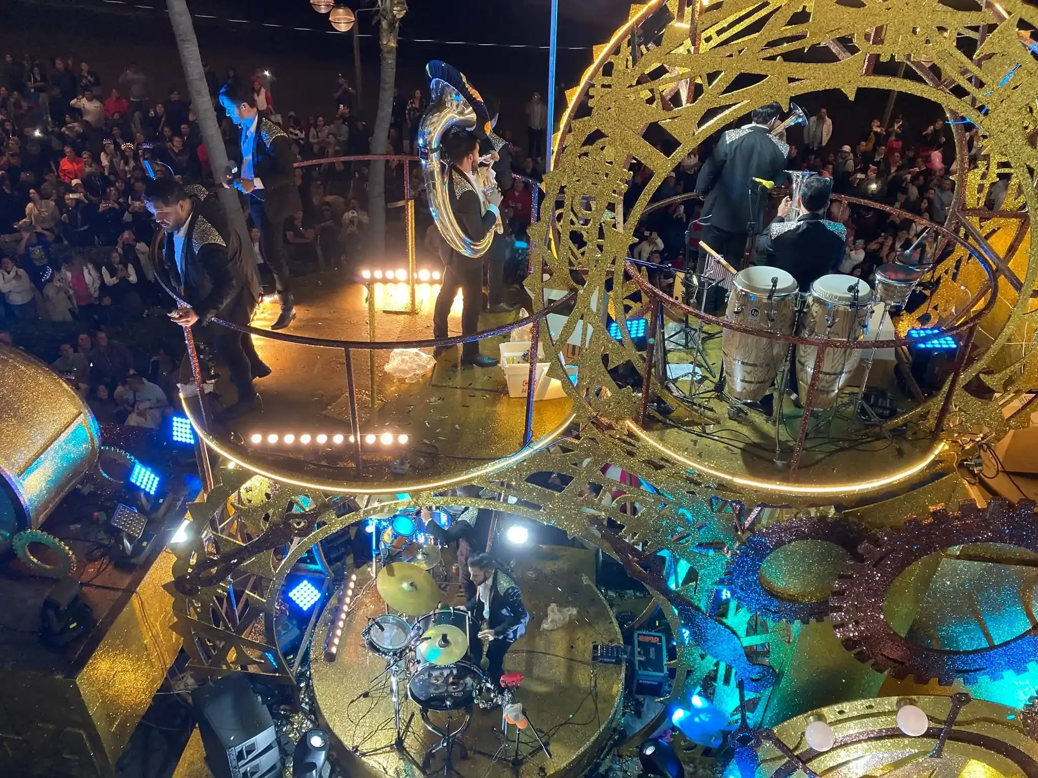 A banda performs in formal sequined jackets on an illuminated gold and blue float, surrounded by a dense crowd along the Mazatlán Malecón at night.