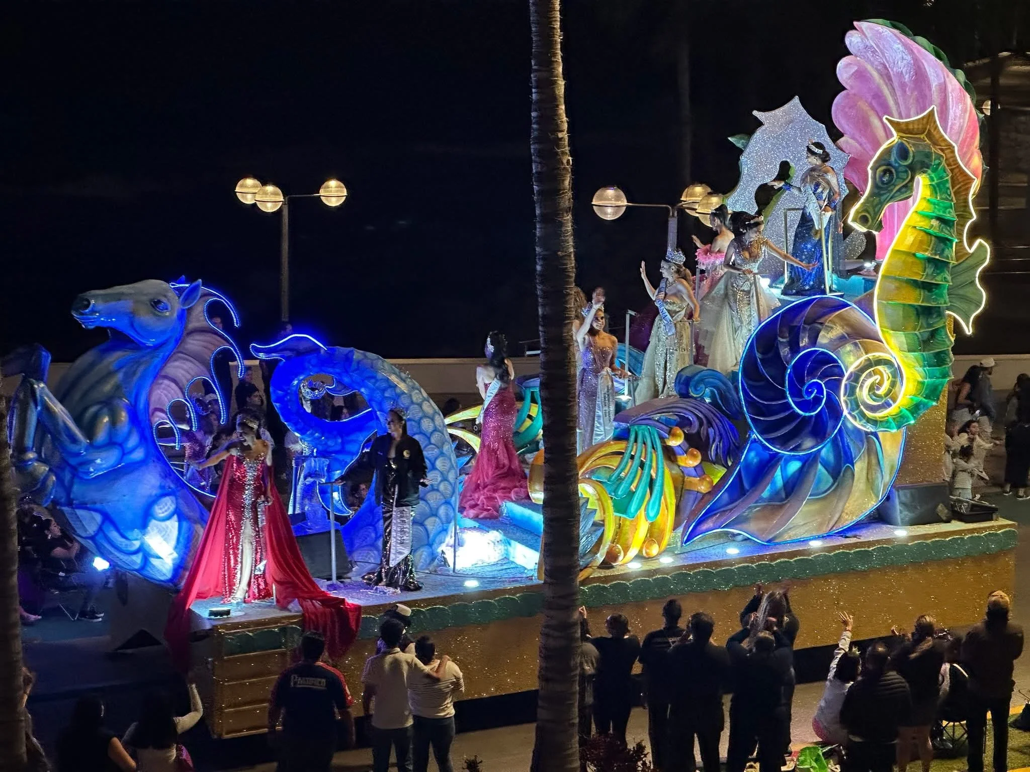 Carnival queens in sequined gowns wave from an illuminated parade float decorated with a giant seahorse, blue horse heads, and ocean-themed sculptures along the Mazatlán Malecón.