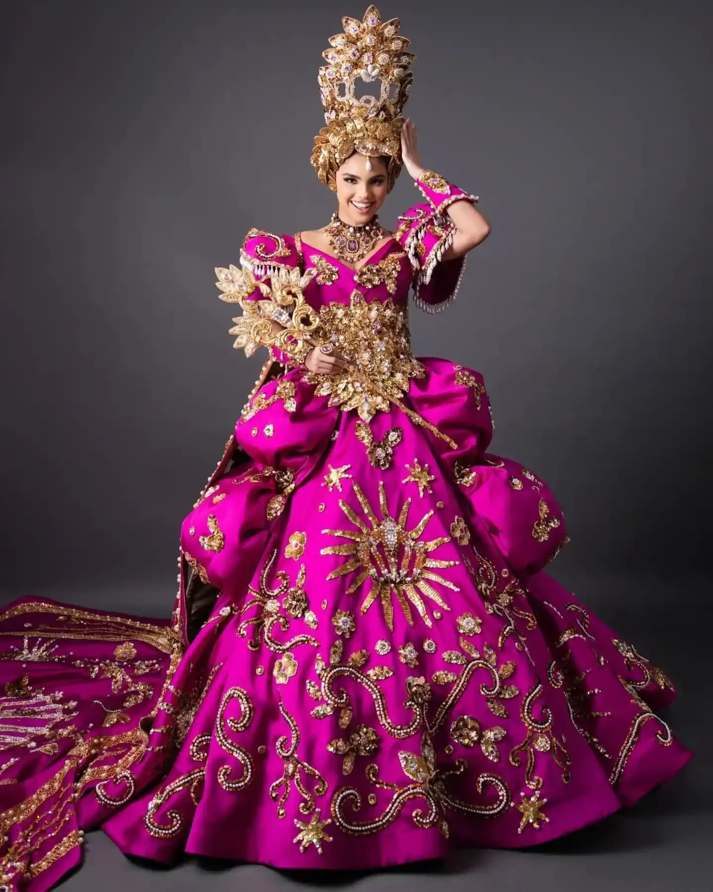 The Queen of Carnaval de Mazatlán in a fuchsia and gold beaded gown with a tall crowned headpiece, posed in studio against a grey backdrop.