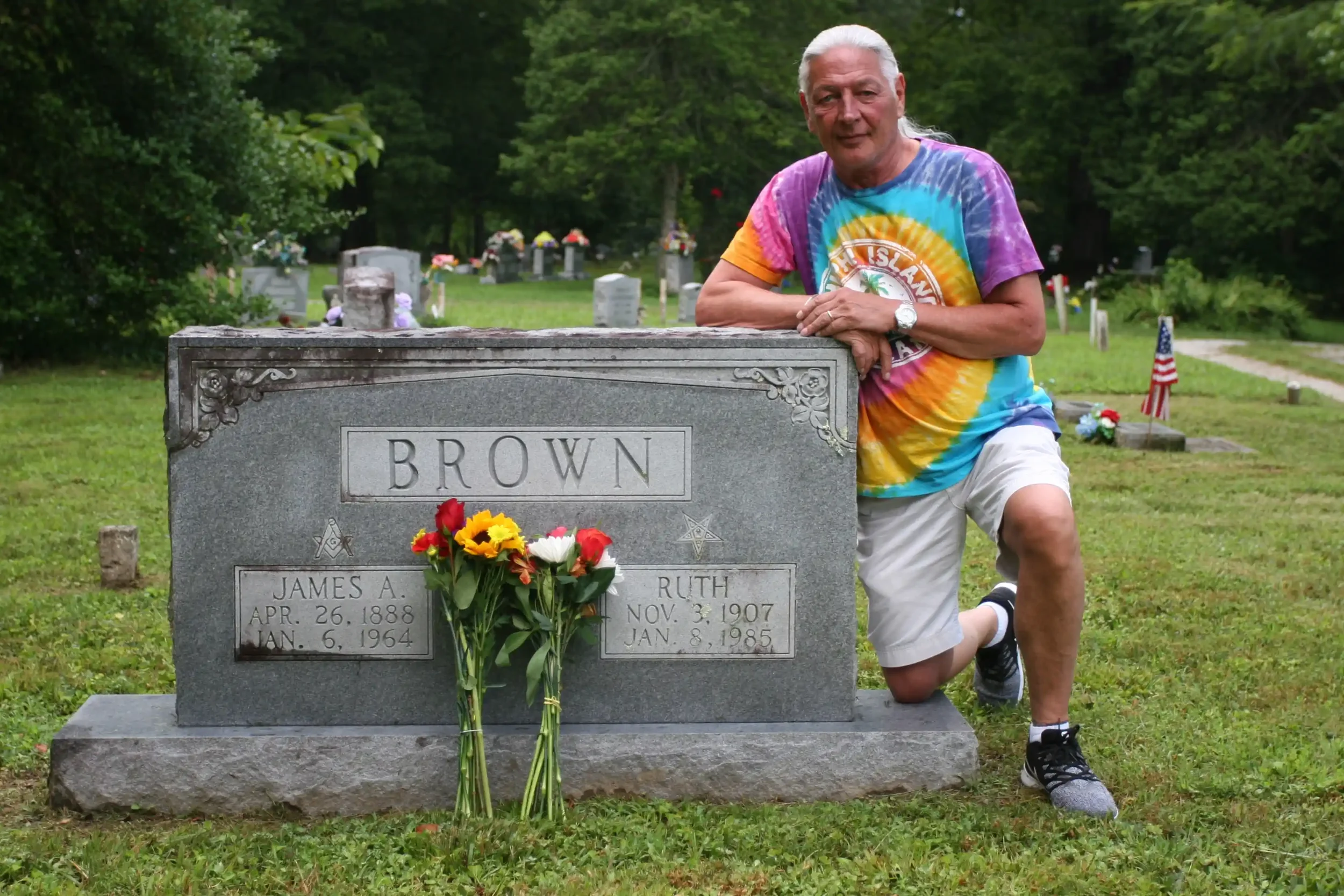 Randy kneels beside the headstone of his grandparents James and Ruth Brown in a Tennessee cemetery, fresh sunflowers and roses propped against the granite.