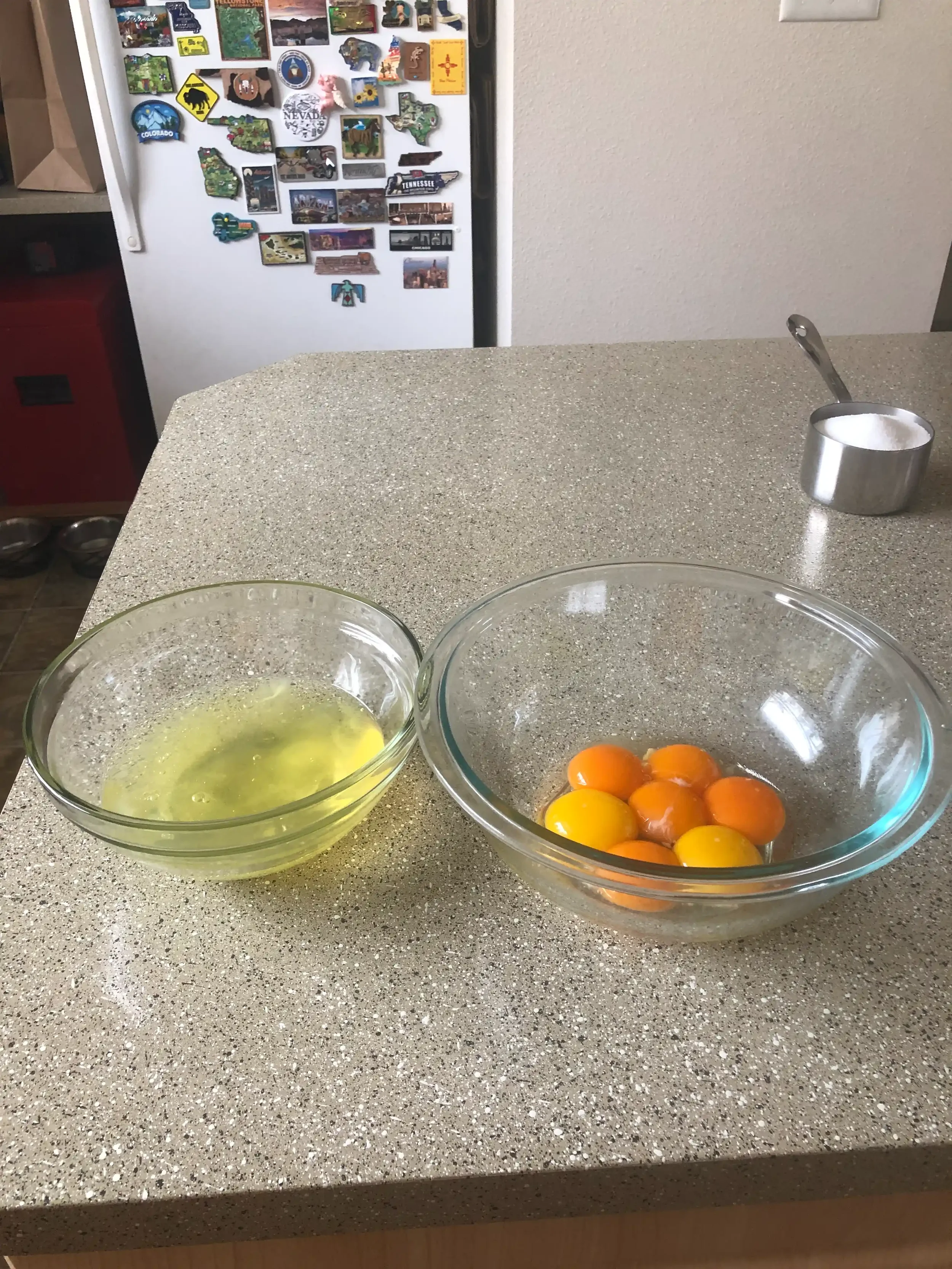 Six egg yolks (with one surprise seventh from a double yolk) separated into a glass bowl beside the bowl of whites, a measuring cup of sugar in the background.