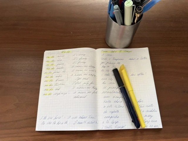 Open notebook with handwritten Spanish vocabulary and sentences, pens beside it on a wooden desk