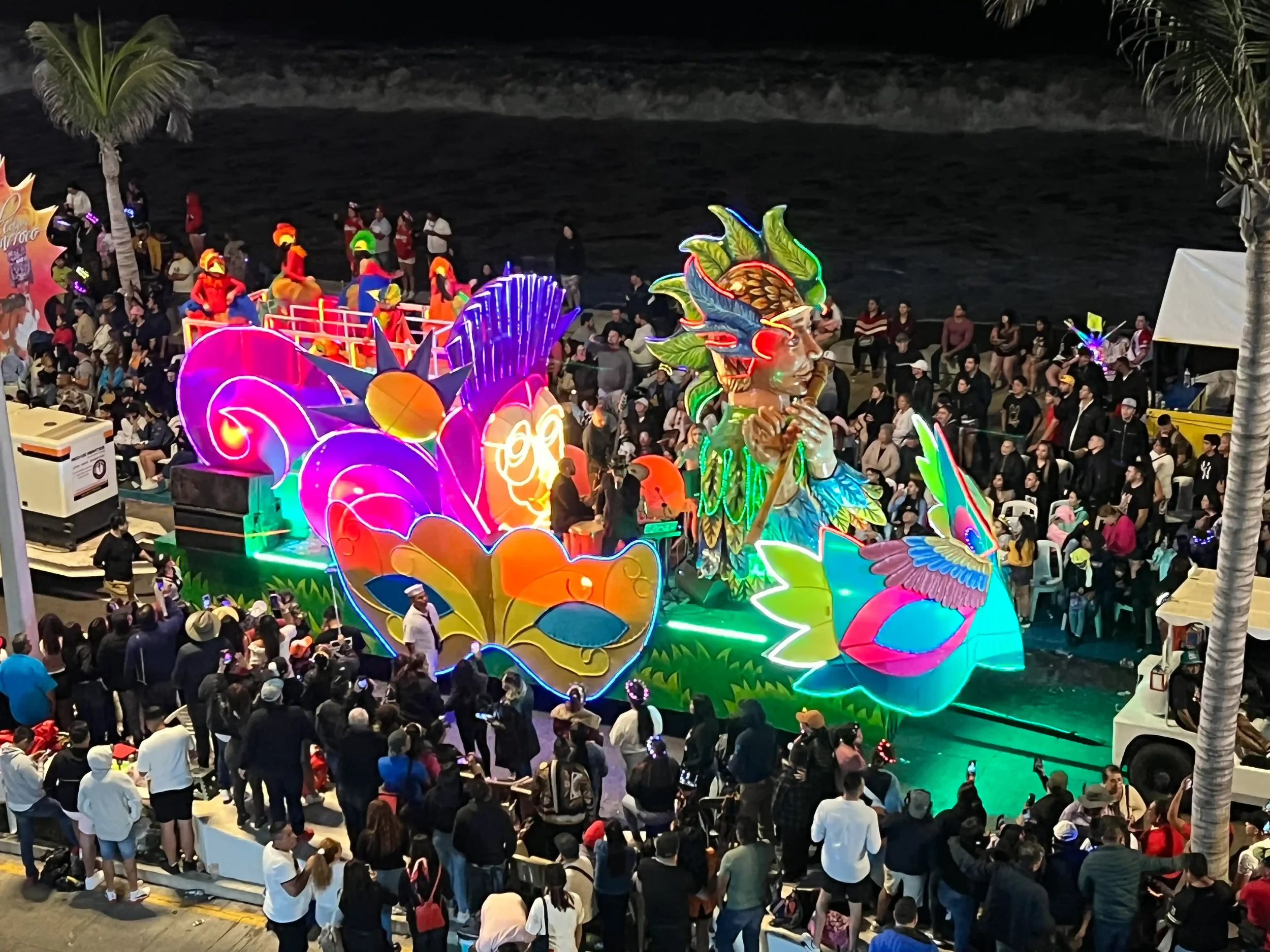 A neon-lit parade float at Carnaval de Mazatlán featuring an oversized masquerade mask and a feathered indigenous figure, with the Pacific Ocean dark behind the Malecón crowd.