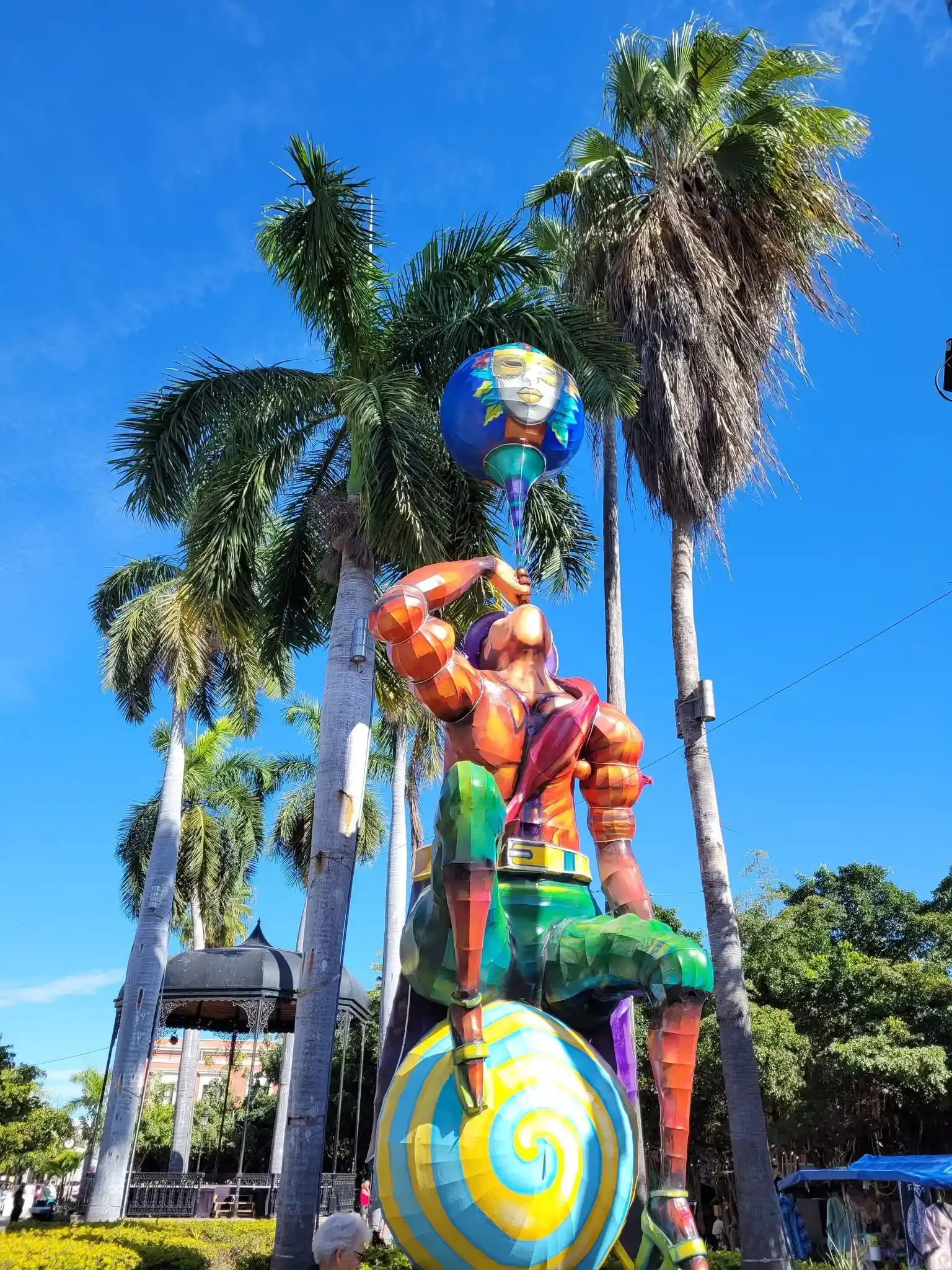 A daytime monigote at Carnaval de Mazatlán, a tall papier-mâché figure of a muscular rainbow-painted man holding a blue mask aloft, standing among palm trees in Plaza Machado.