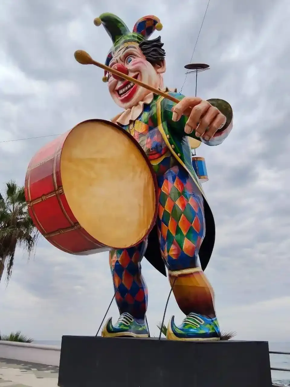 A Carnaval de Mazatlán monigote of a harlequin clown in a checkered diamond-pattern suit, holding a large red and tan tambora drum mid-beat, photographed from below against an overcast sky.