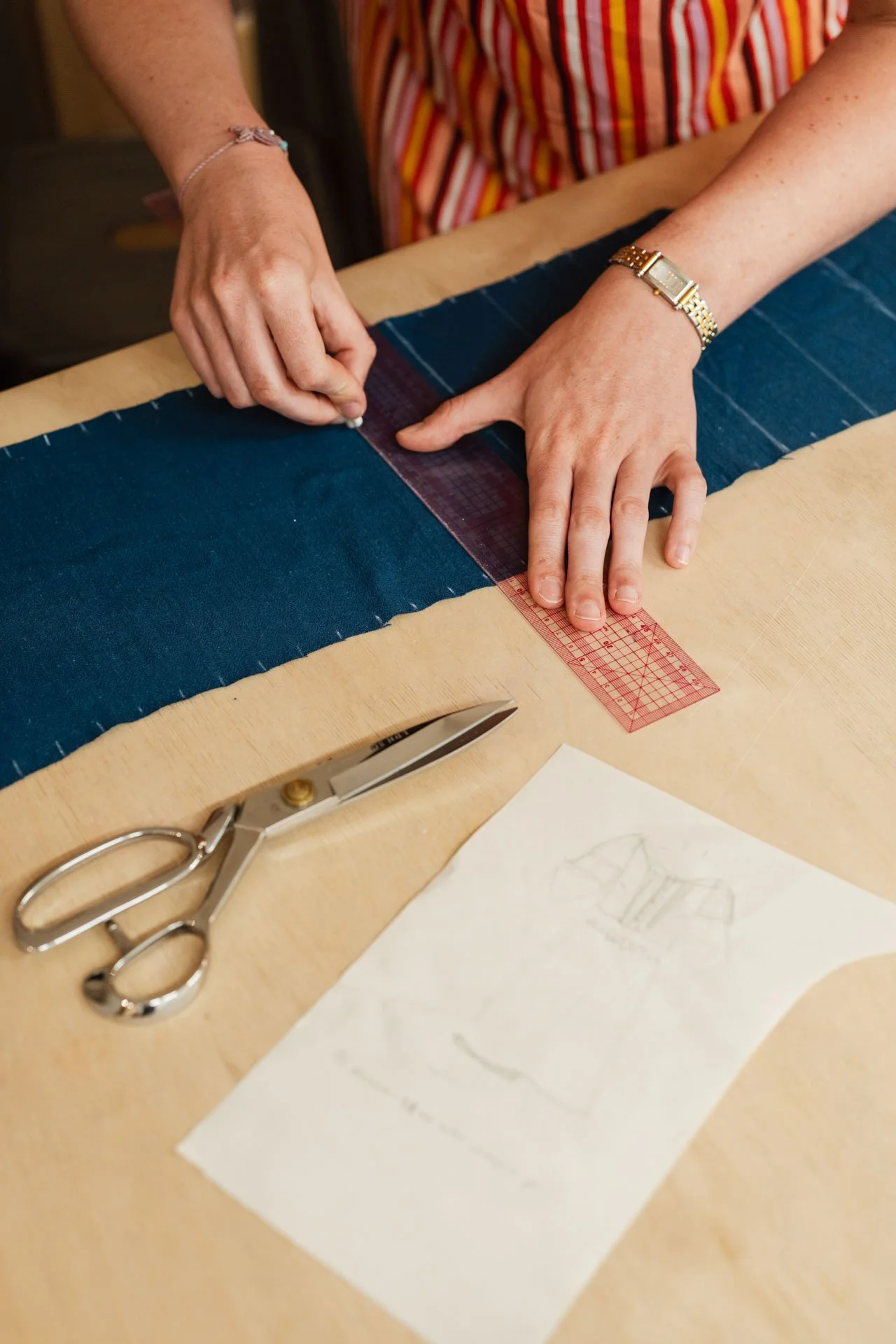 Une personne mesure du tissu avec une règle en plastique rouge sur une table en bois, avec des ciseaux en métal et un dessin esquissé d'un vêtement.