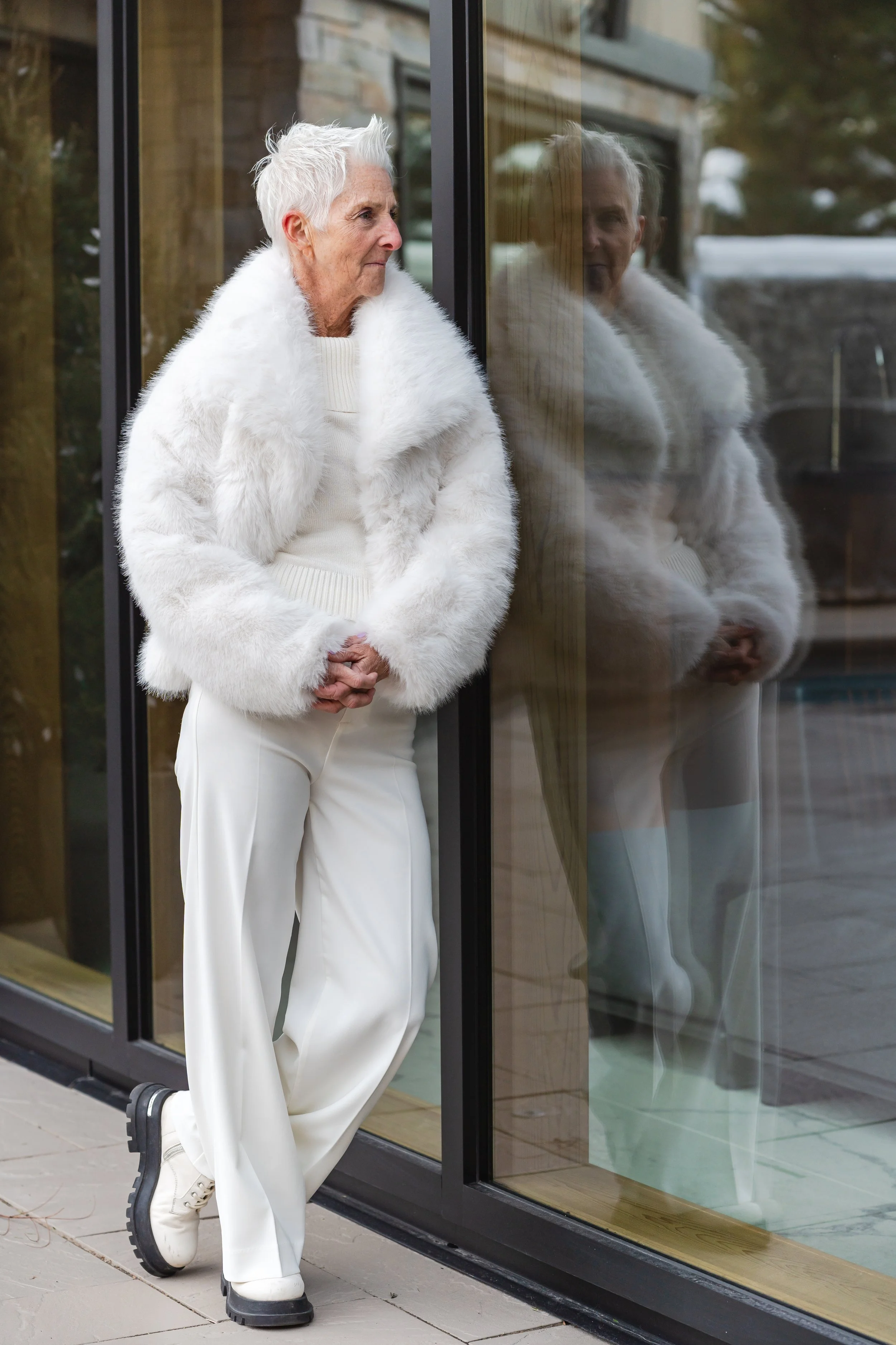 Une femme âgée aux cheveux courts gris, portant un manteau blanc en peluche, un pull blanc, et un pantalon blanc, se tient contre une vitre reflet. Elle regarde pensivement à l'extérieur.