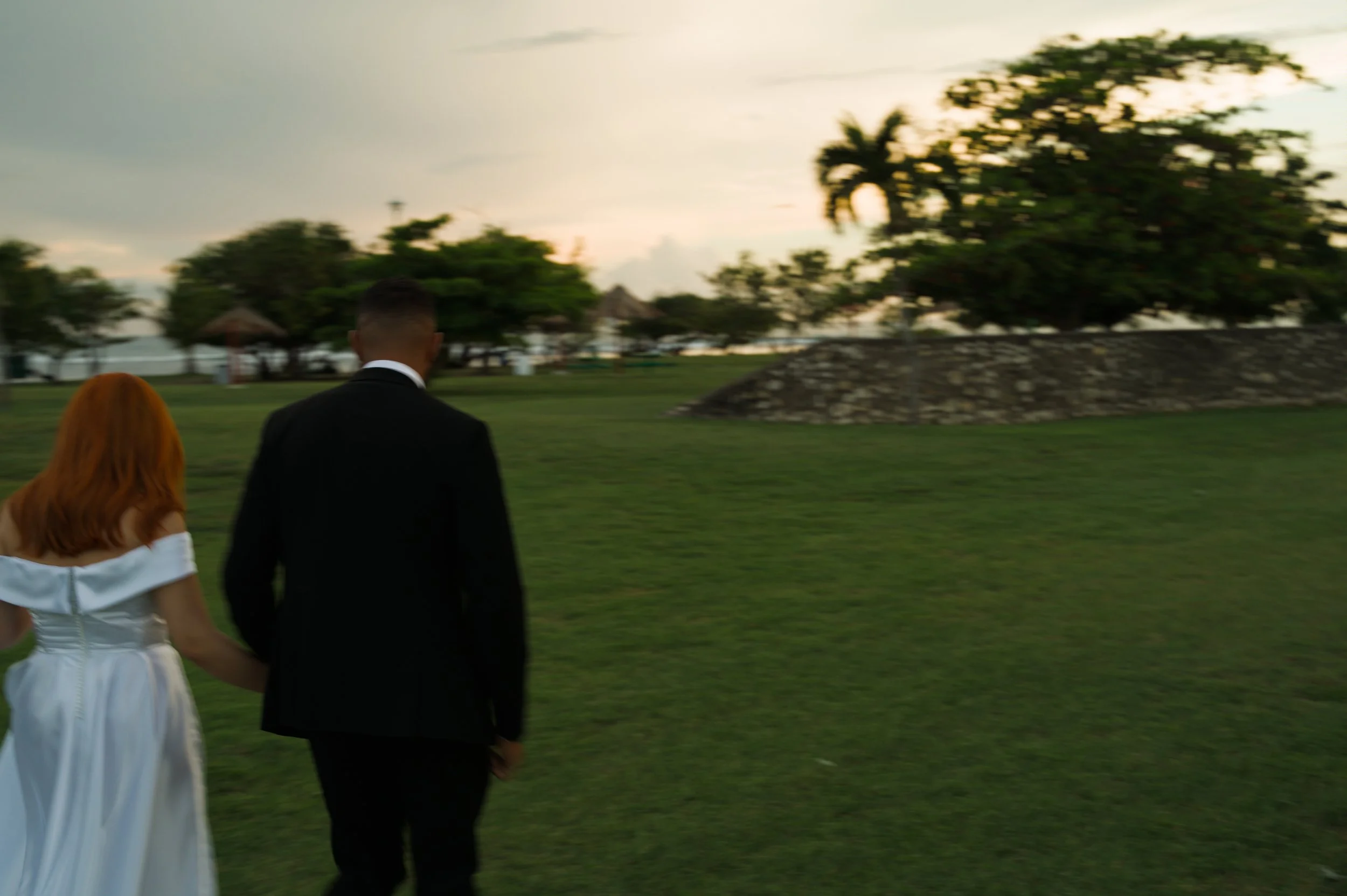 A couple is walking away from the camera on a grassy lawn during sunset. The woman has red hair and is wearing a white dress, and the man is wearing a black suit.
