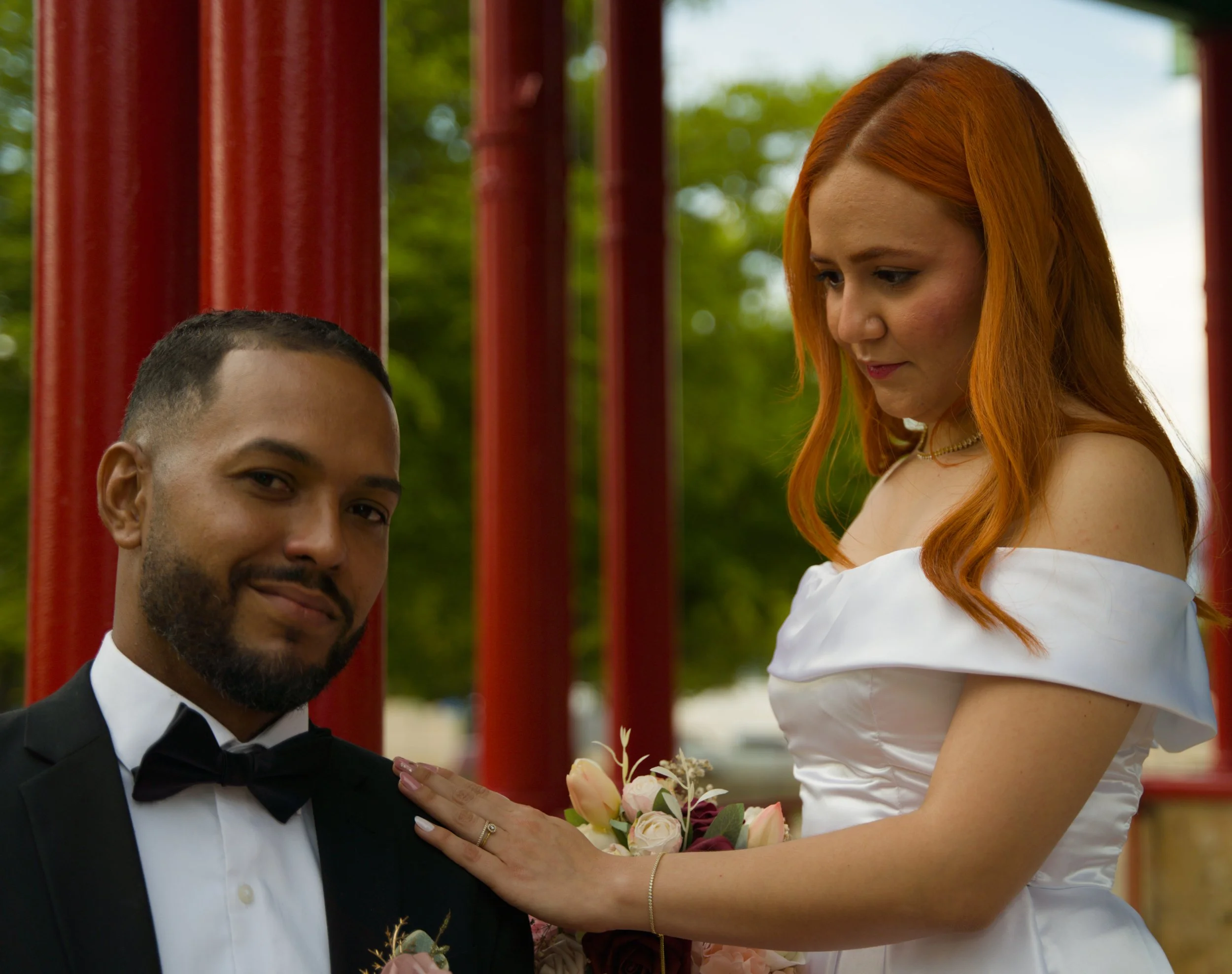 A bride and groom pose outdoors, with the bride placing her hand on the groom's shoulder, both smiling softly. The groom is in a black tuxedo with a bow tie, and the bride is wearing an off-shoulder white wedding dress, holding a bouquet of flowers. 
