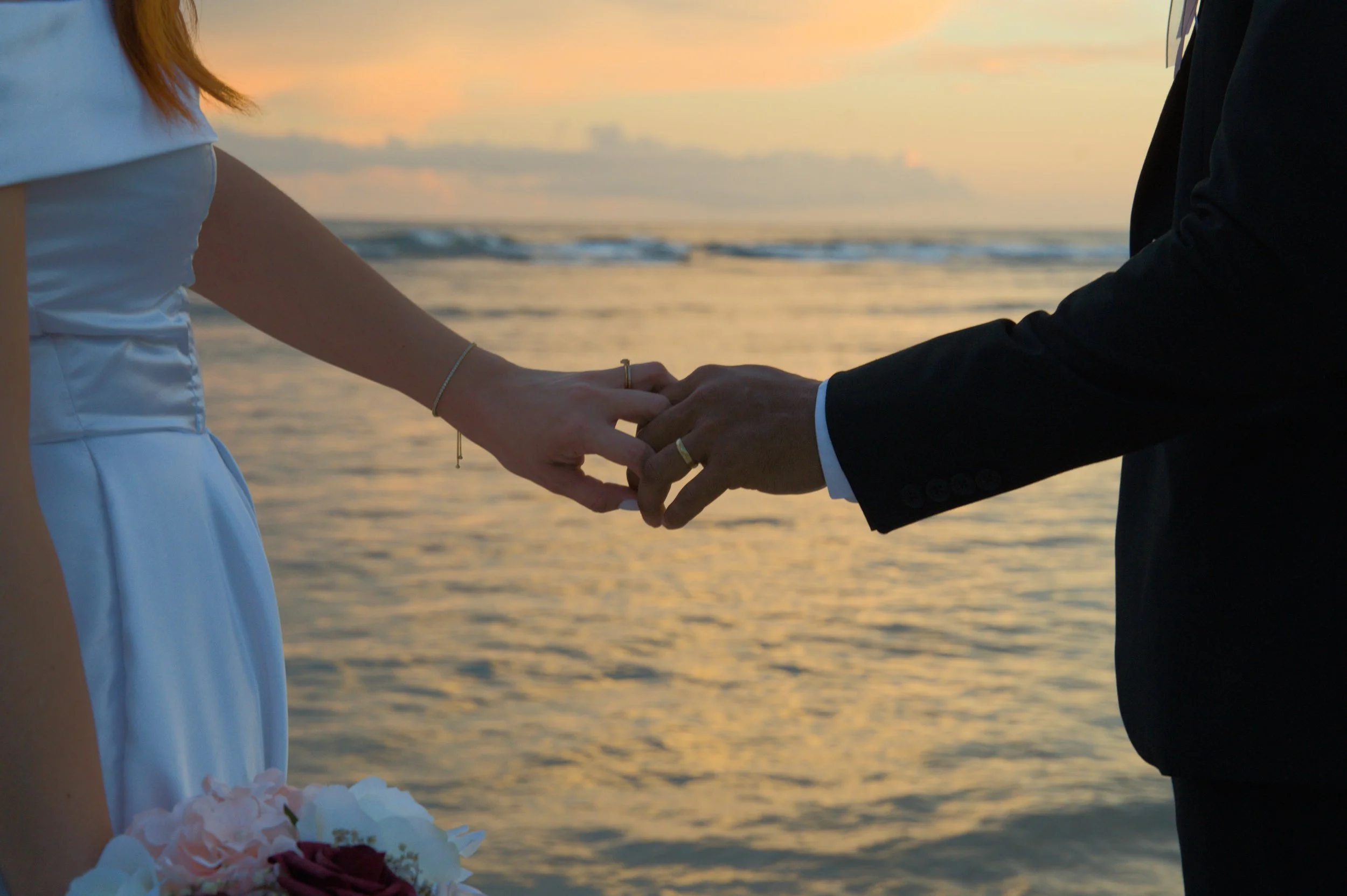 Close-up of a couple holding hands during a sunset at the beach, with the bride holding a bouquet of flowers.