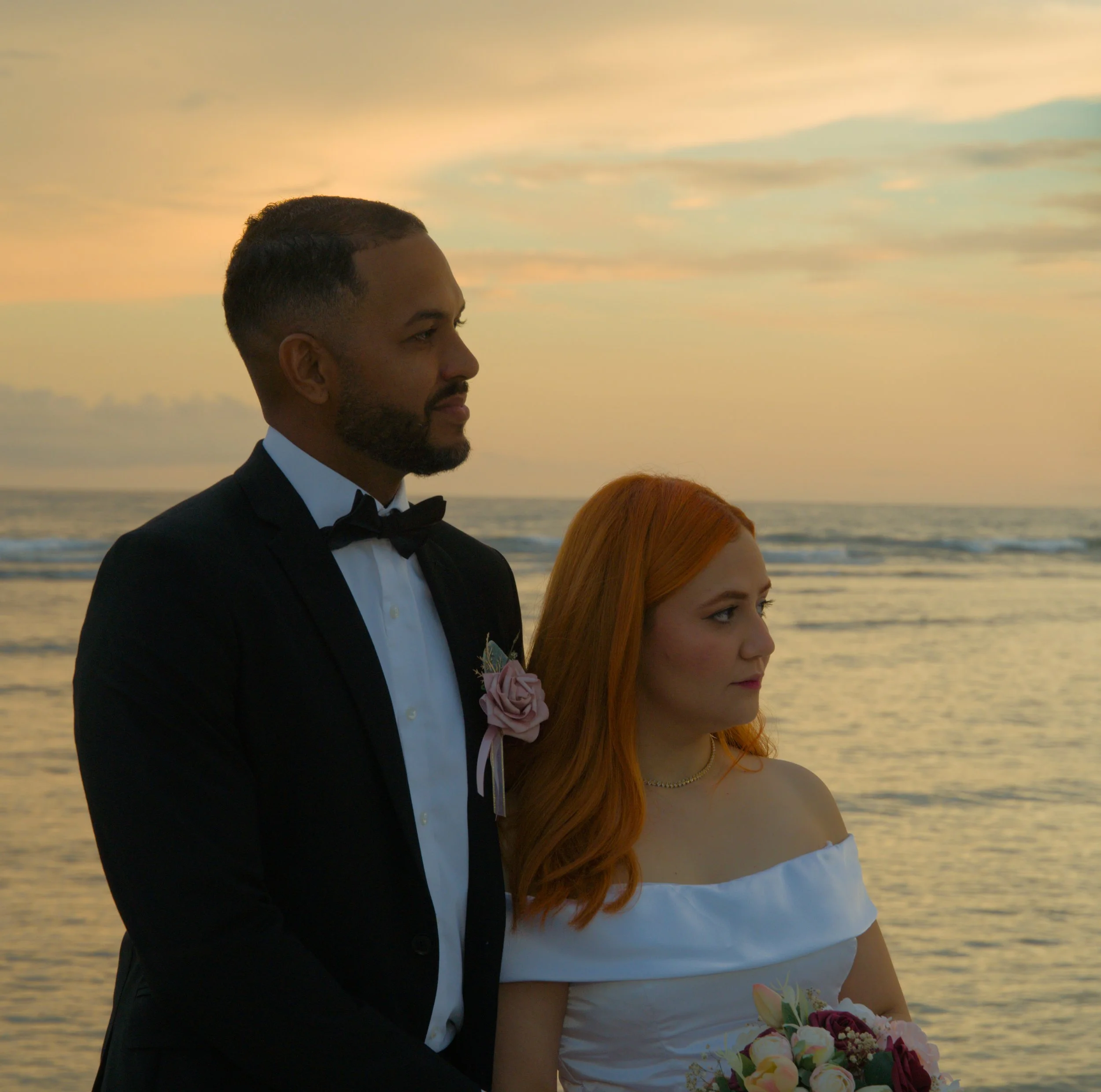 A couple in wedding attire standing on the beach at sunset, facing opposite directions with calm ocean waves in the background.