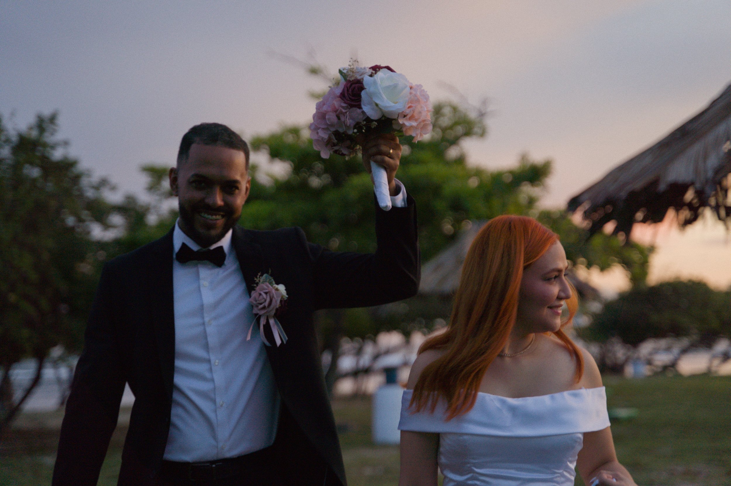 A newlywed couple outdoors at sunset in Puerto Rico, the groom holding a bouquet of flowers high, smiling, and the bride with red hair in an off-shoulder white dress, looking away, with trees and a rustic thatched umbrella in the background.