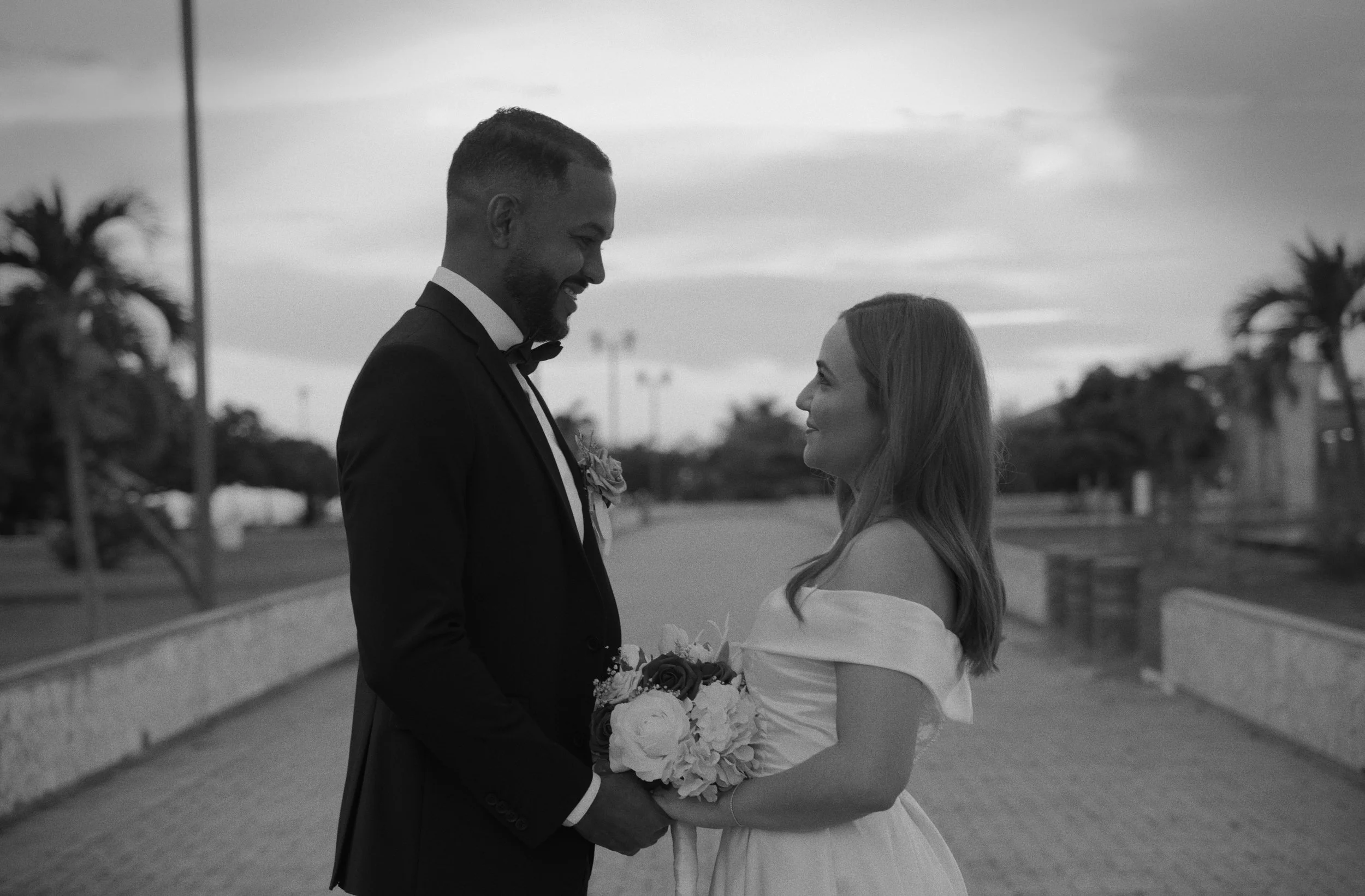A black-and-white photo of a bride and groom standing face to face outdoors, smiling at each other, with the bride holding a bouquet of flowers.