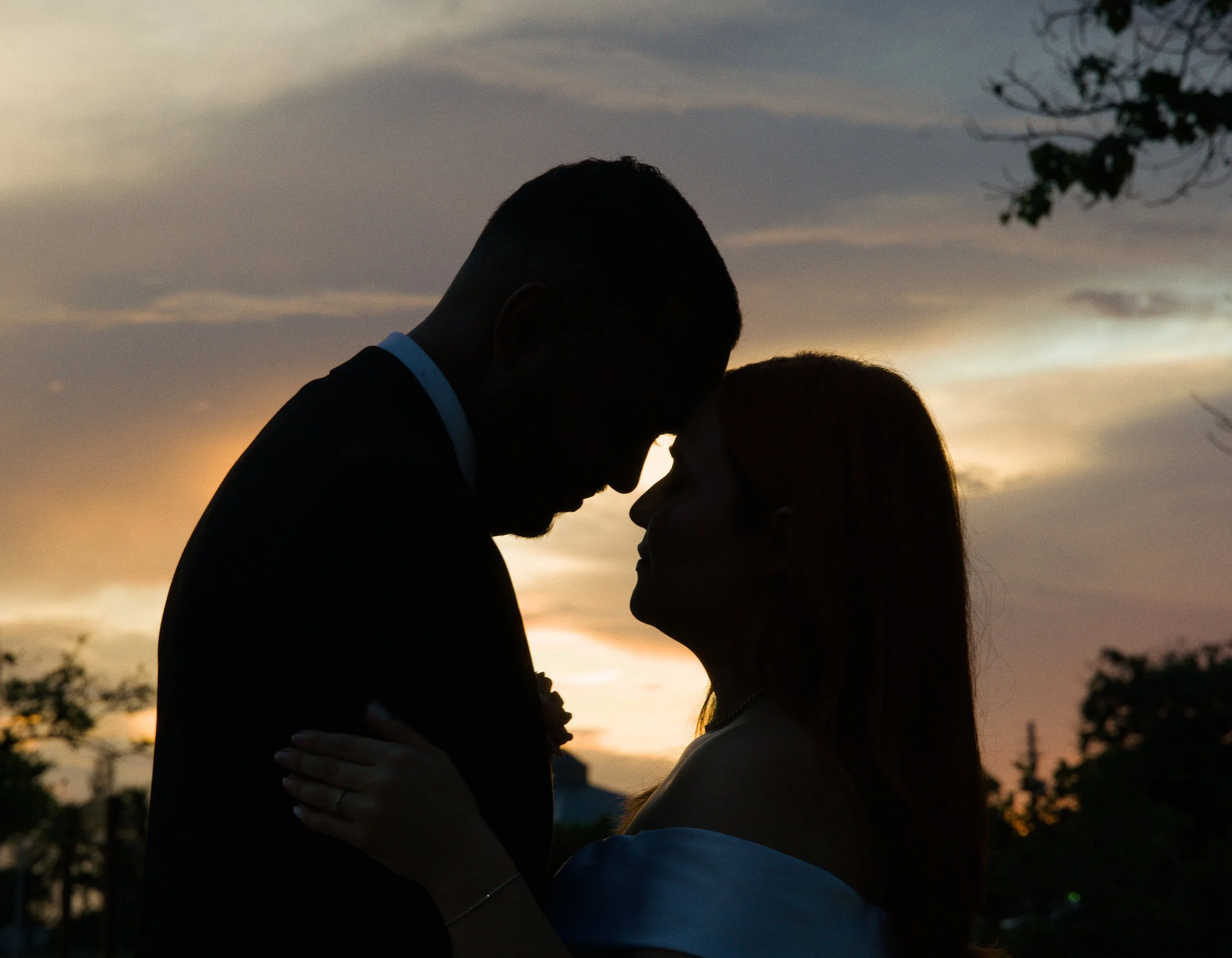 Silhouette of a couple facing each other closely at sunset, with the sky lit in warm colors and trees in the background.