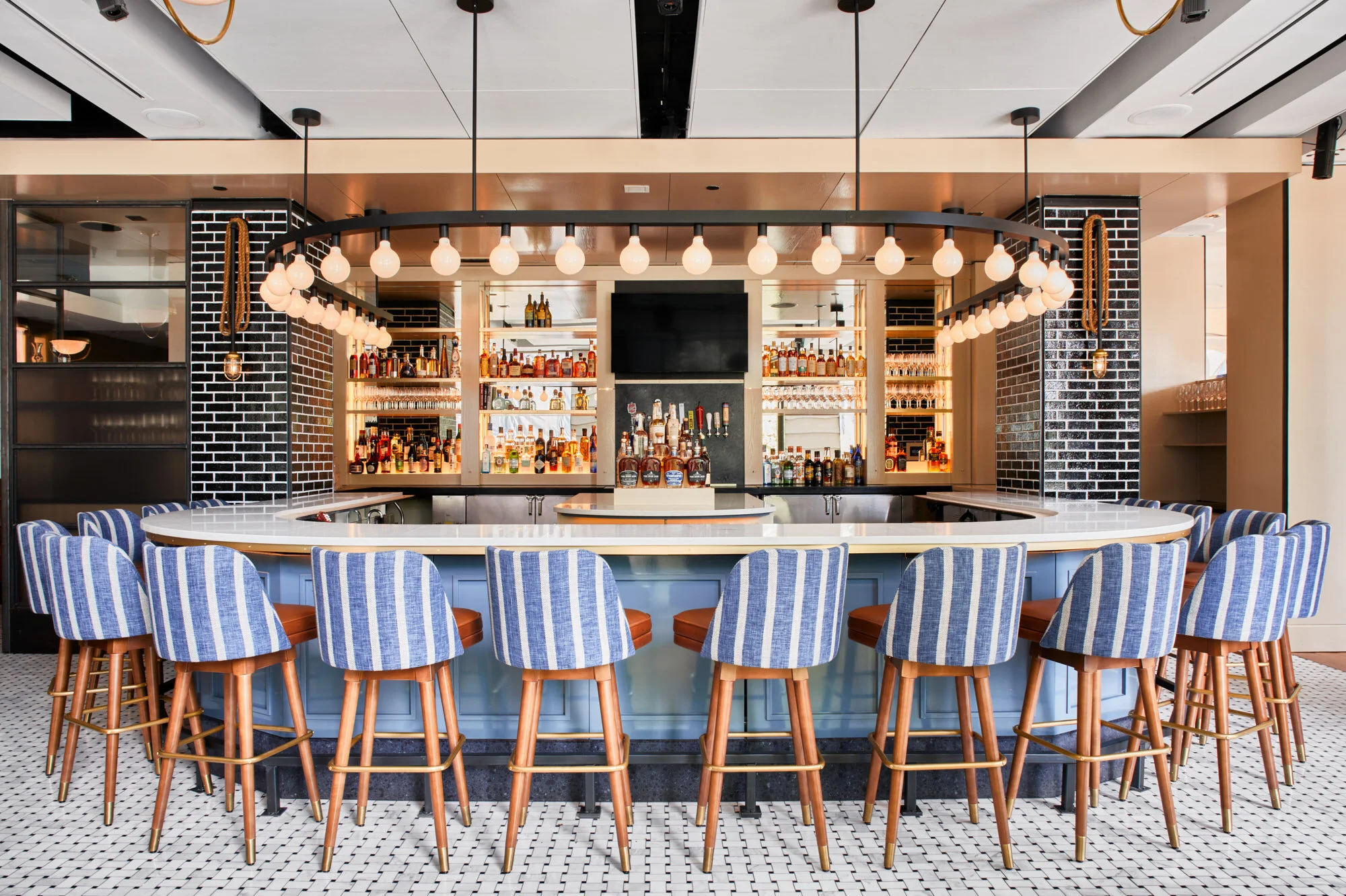 A bar with a curved white countertop, surrounded by wooden stools with blue and white striped upholstery. Behind the bar, there are shelves stocked with various bottles of alcohol, glassware, and a black television screen. The bar area features industrial-style decor with black brick accents, pendant lighting, and a ceiling with modern paneling and exposed lighting fixtures. The floor has a black and white tiled pattern.