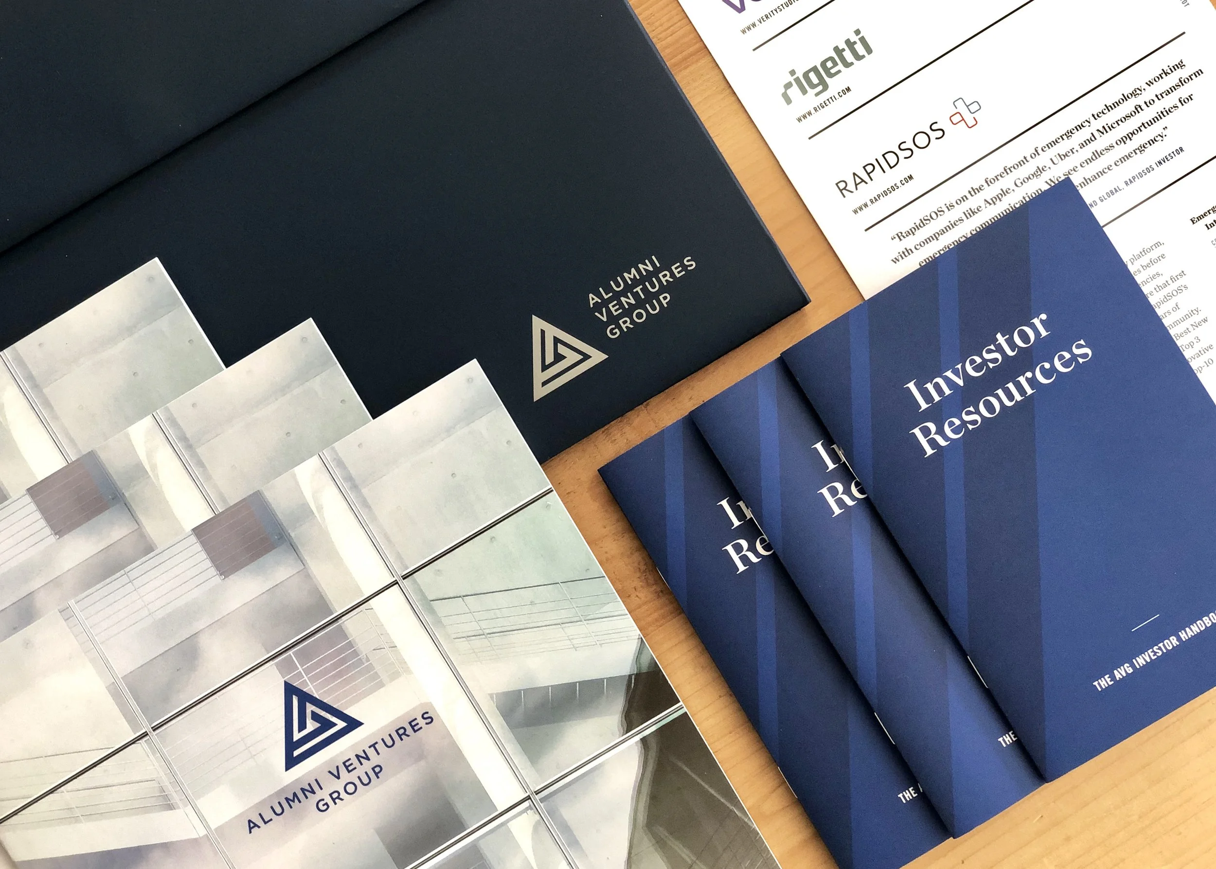 Various printed materials including brochures and pamphlets on a wooden surface. The brochures display titles such as 'ALUMNI VENTURES GROUP,' 'Investing Resources,' and 'RapidsOS.' Some printed pages also show color samples or swatches.