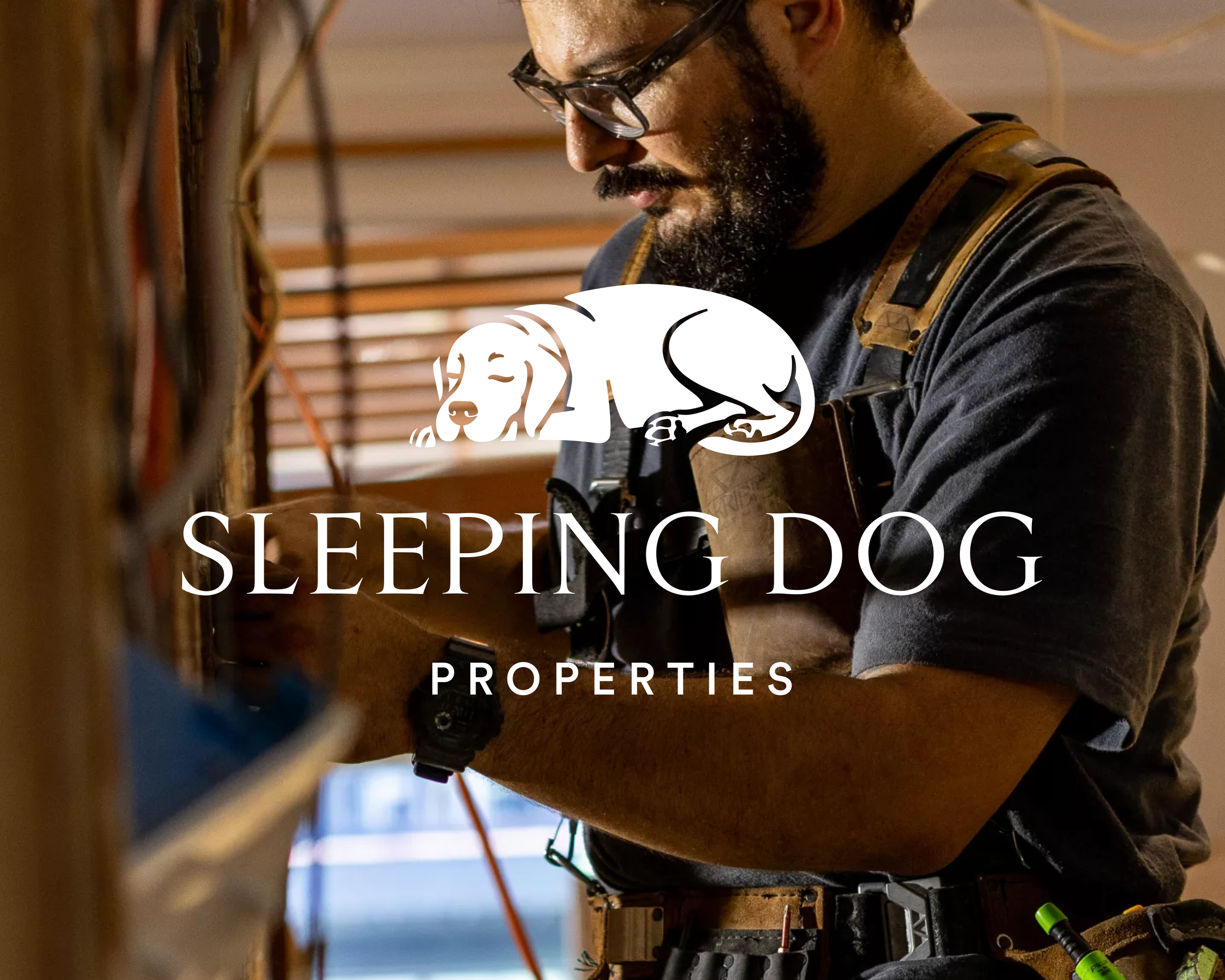 A man with glasses and a beard focuses on woodworking, working on a wooden surface with visible tools and wires, in a workshop with wooden beams in the background. The overlay text reads 'Sleeping Dog Properties'.