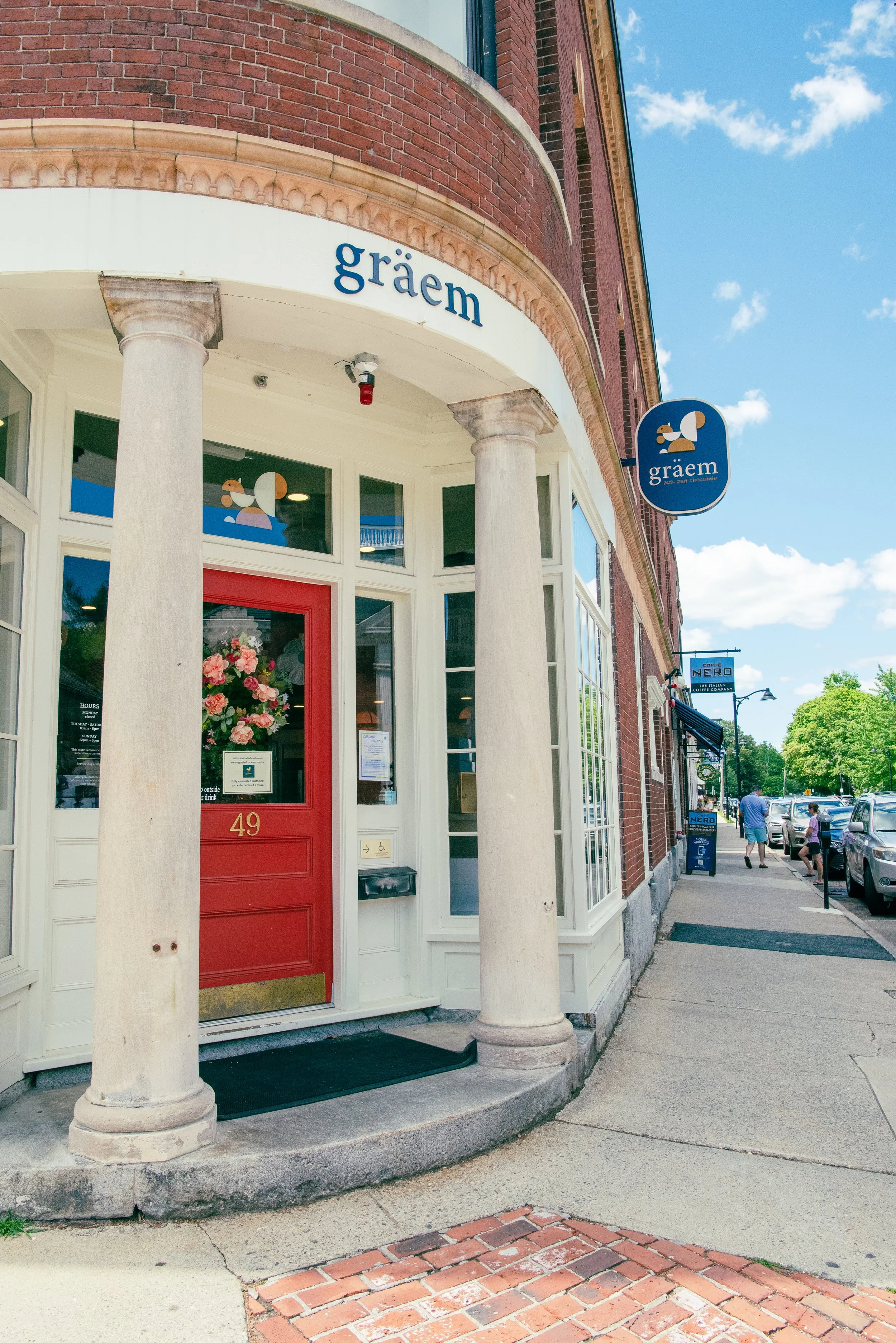 Facade of a storefront named 'gräem' with a red door, white columns, and a blue sign, on a sunny day with a brick sidewalk and pedestrians.
