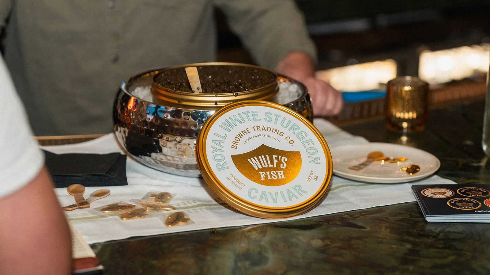 A container of Wulf's Fish caviar on a table, with a spoon inside and small samples of caviar on a plate nearby, with other items and a person in the background.
