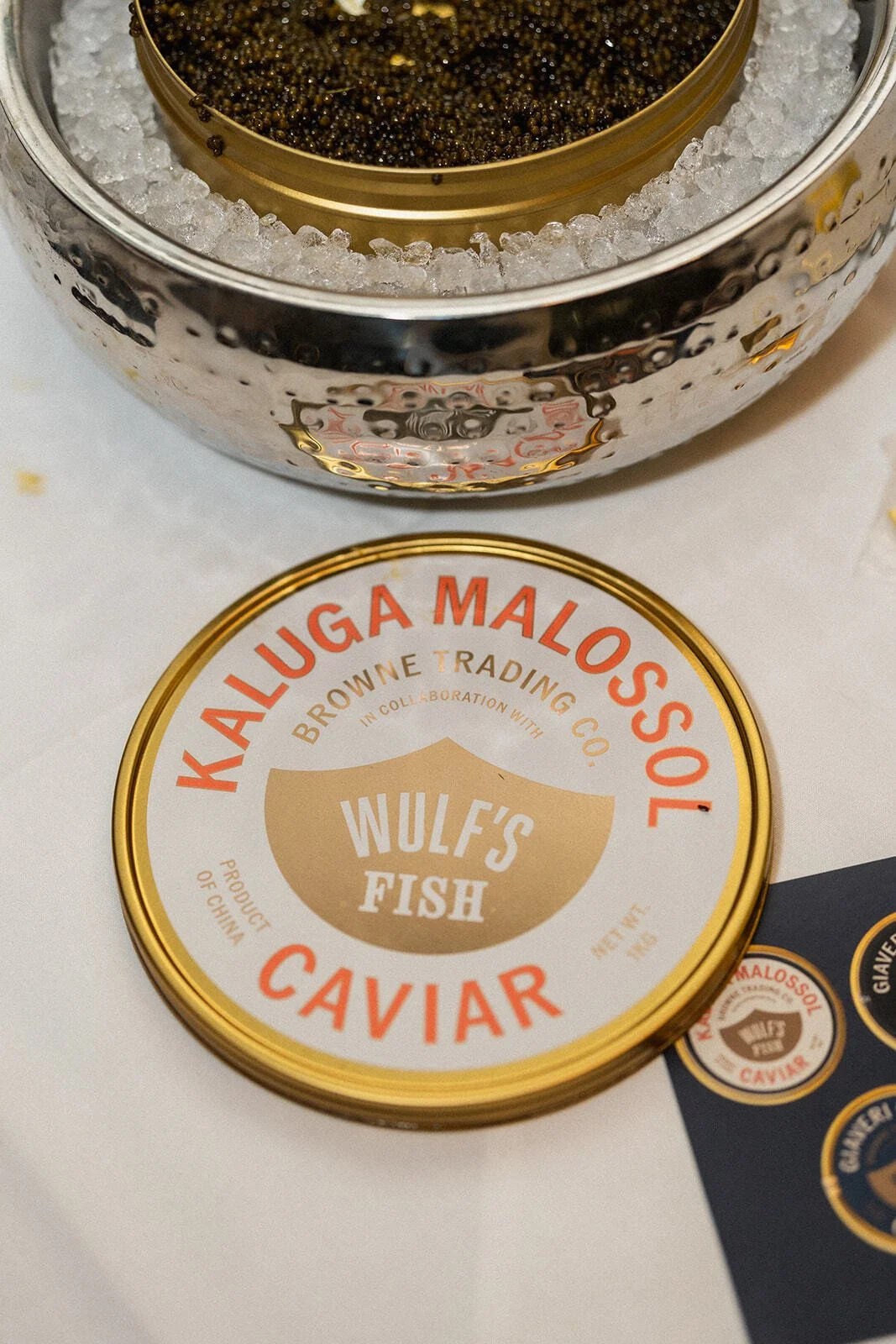 A tin of Wulf's Fish Caviar from Kaluga Malossol with black fish eggs inside, placed next to a metallic container filled with ice.