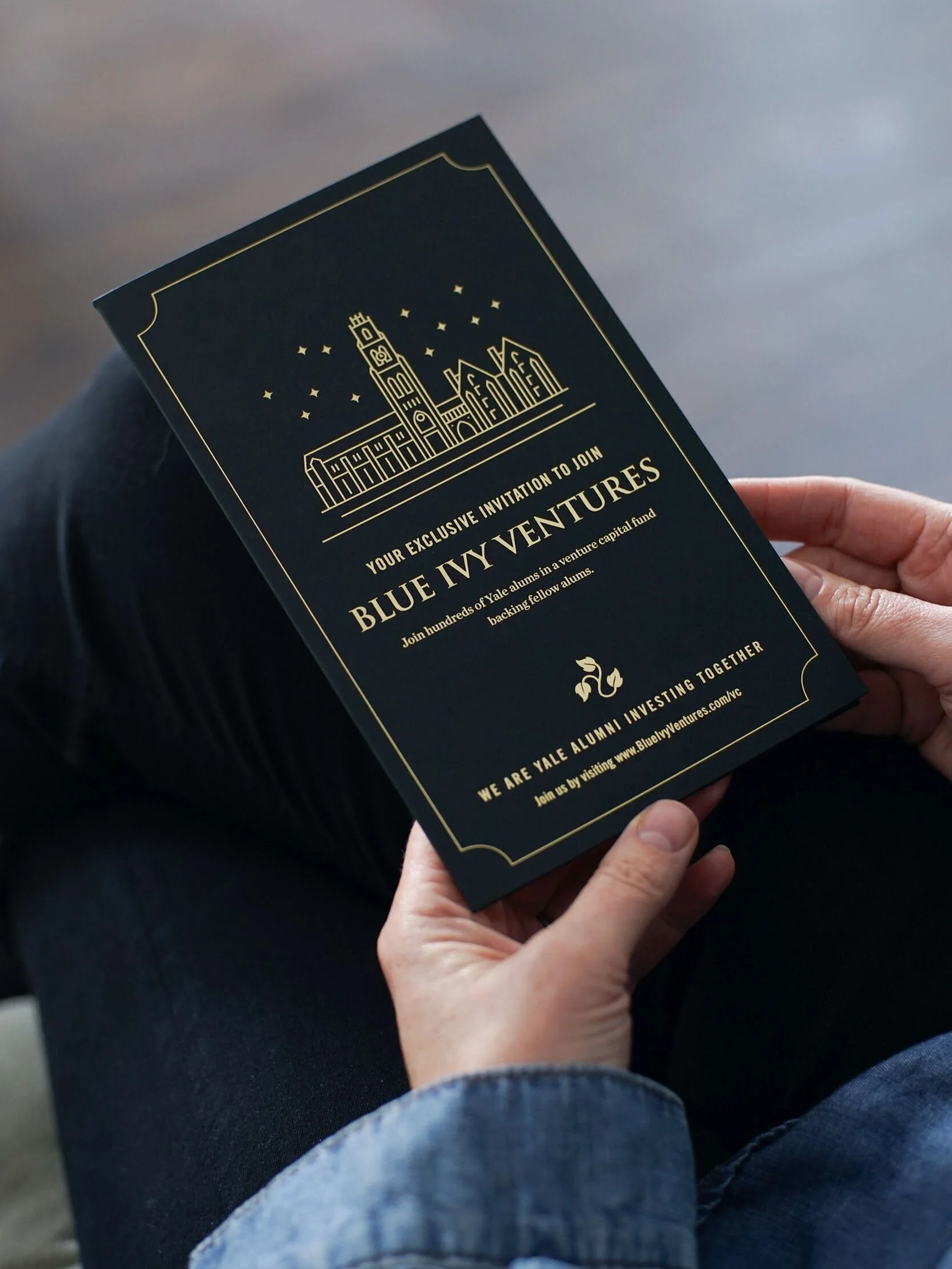 A person holding a black invitation card with gold detailing, featuring an illustration of a clock tower and buildings, promoting an exclusive Yale alumni venture capital fund event.