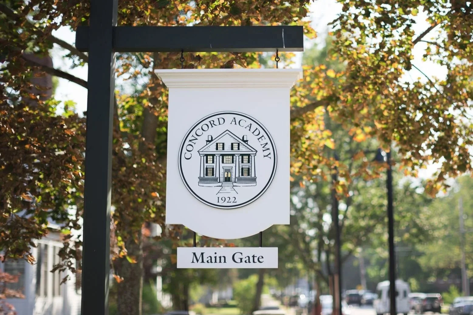 A white sign with a crest displaying a house and the words 'Concord Academy 1922', with a smaller sign reading 'Main Gate' hanging below area.