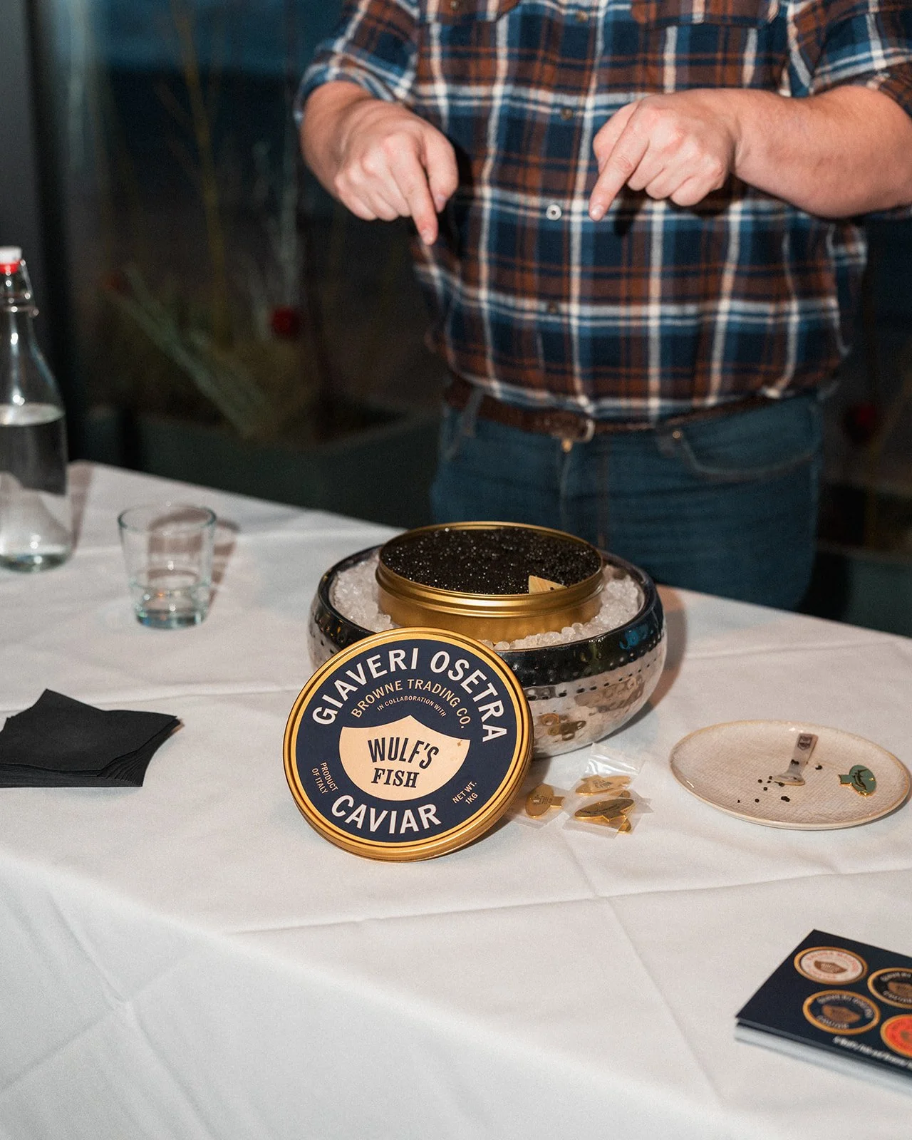 A person in a plaid shirt is pointing down at a container of black caviar labeled "Wulf's Fish" from Giaveri Osteria, sitting on a table covered with a white tablecloth. There are small plates, glasses, a bottle of water, a spoon, and some coins scattered on the table.