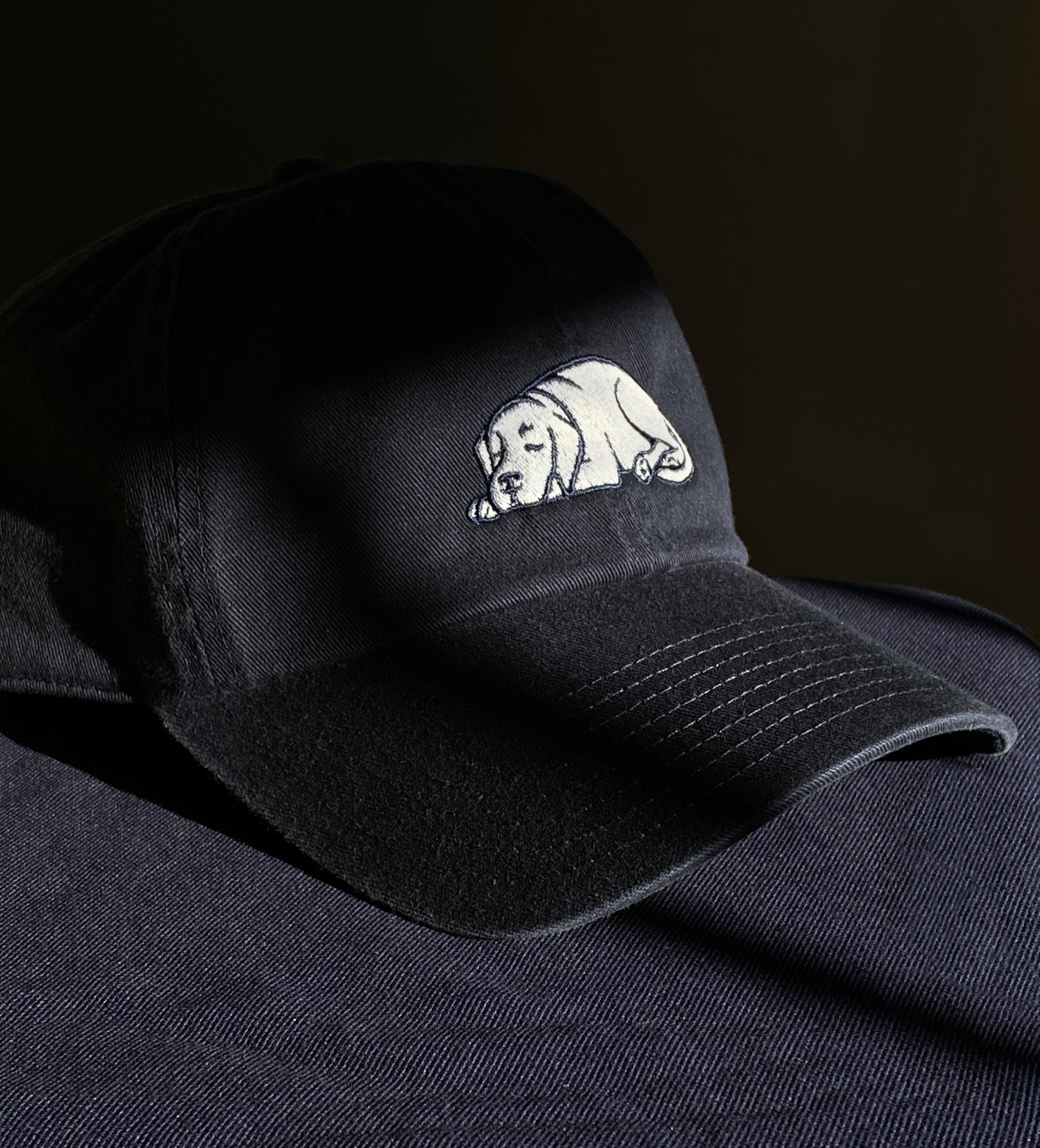 Black baseball cap with a white embroidered logo for 'Sleeping Dog Properties' on the front, placed on a dark fabric surface.