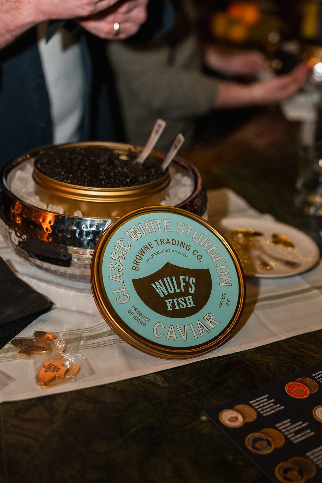 A tin of Wulf's Fish Caviar from Classic White Sturgeon Caviar, displayed on a table with a person in the background taking a photo with a smartphone. The tin is open, revealing black caviar, and is placed on a bed of ice.
