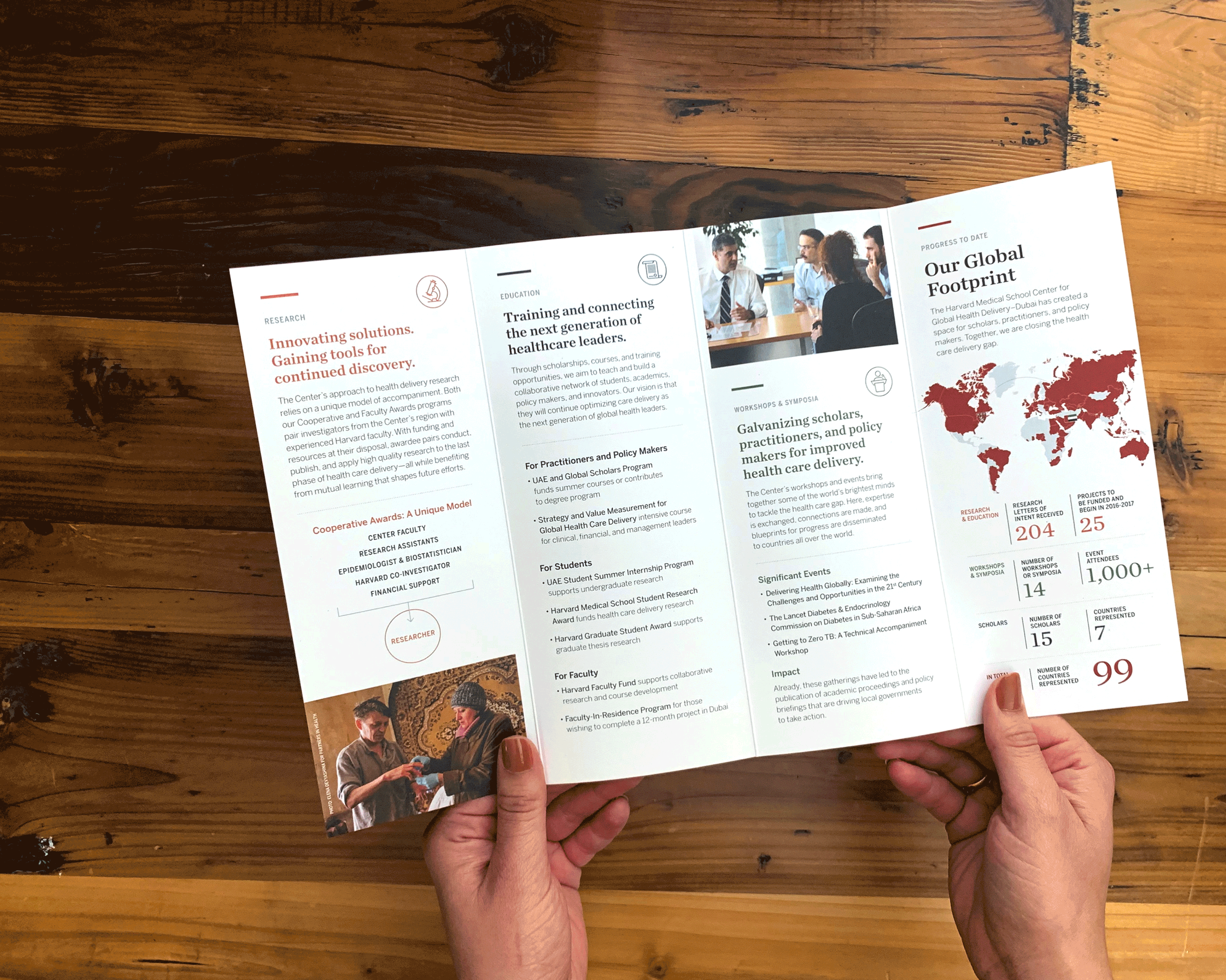 Close-up of a person's hands holding an open brochure or pamphlet on a wooden surface. The brochure features images and text related to healthcare, research, education, and global health initiatives.
