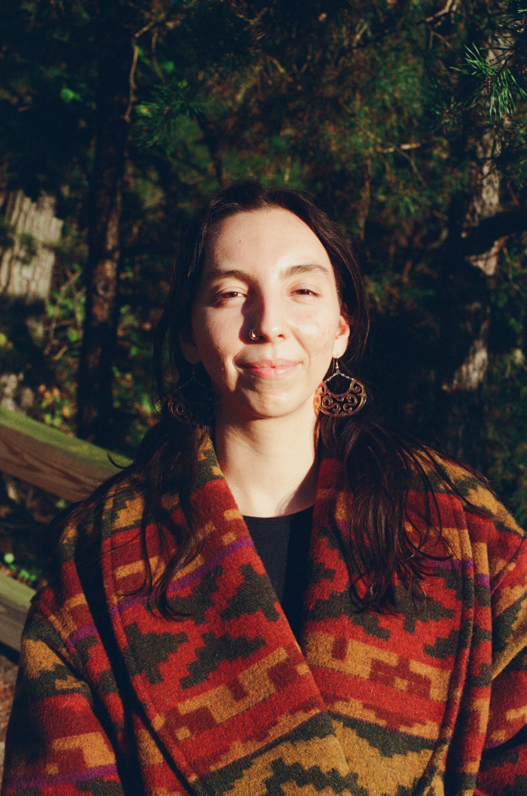 A woman with dark hair, wearing large earrings and a colorful patterned coat, stands outdoors in a sunlit forested area.