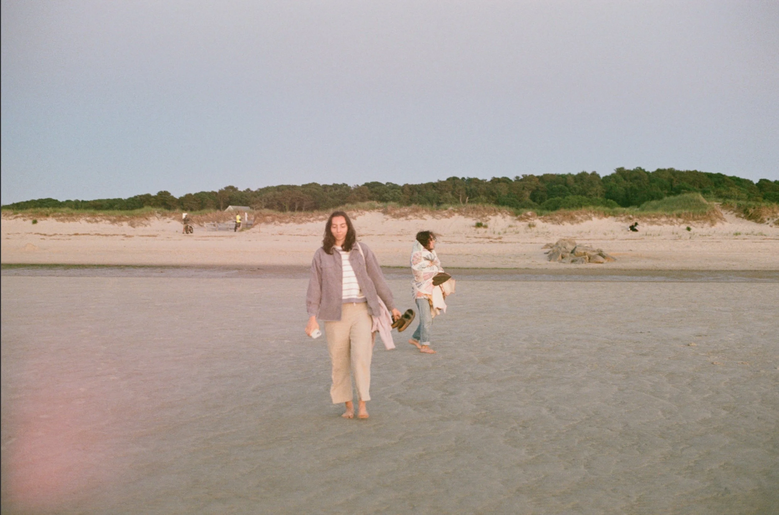 Two women walking on a sandy beach during sunset, with dunes and greenery in the background.