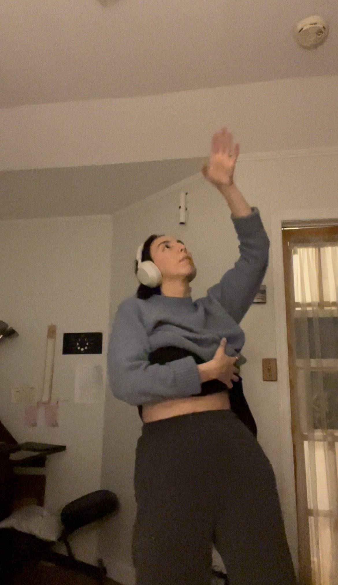 A woman wearing headphones, a gray sweatshirt, and black pants is standing indoors, lifting her shirt to reveal her stomach, and gesturing with her right hand towards the ceiling.