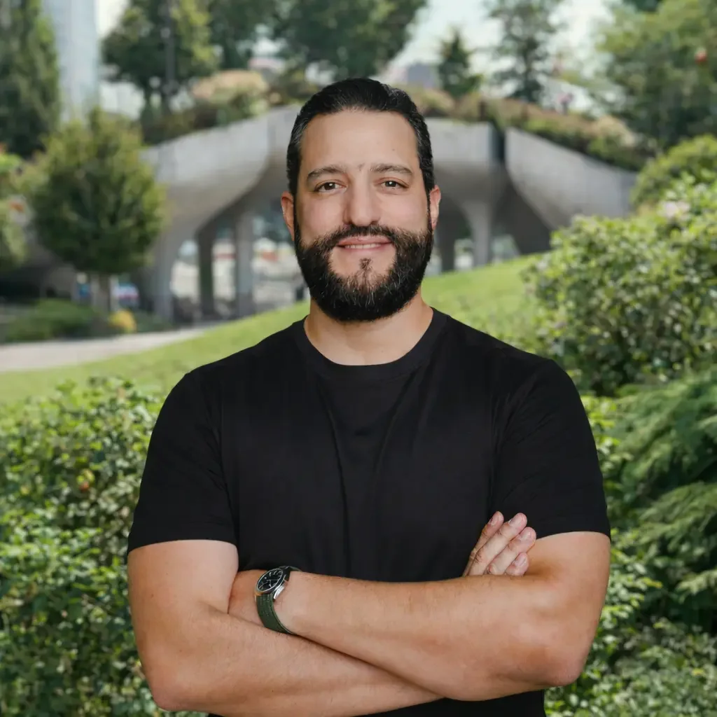 Hombre con barba y camiseta negra sonriendo en un parque con jardines y árboles.