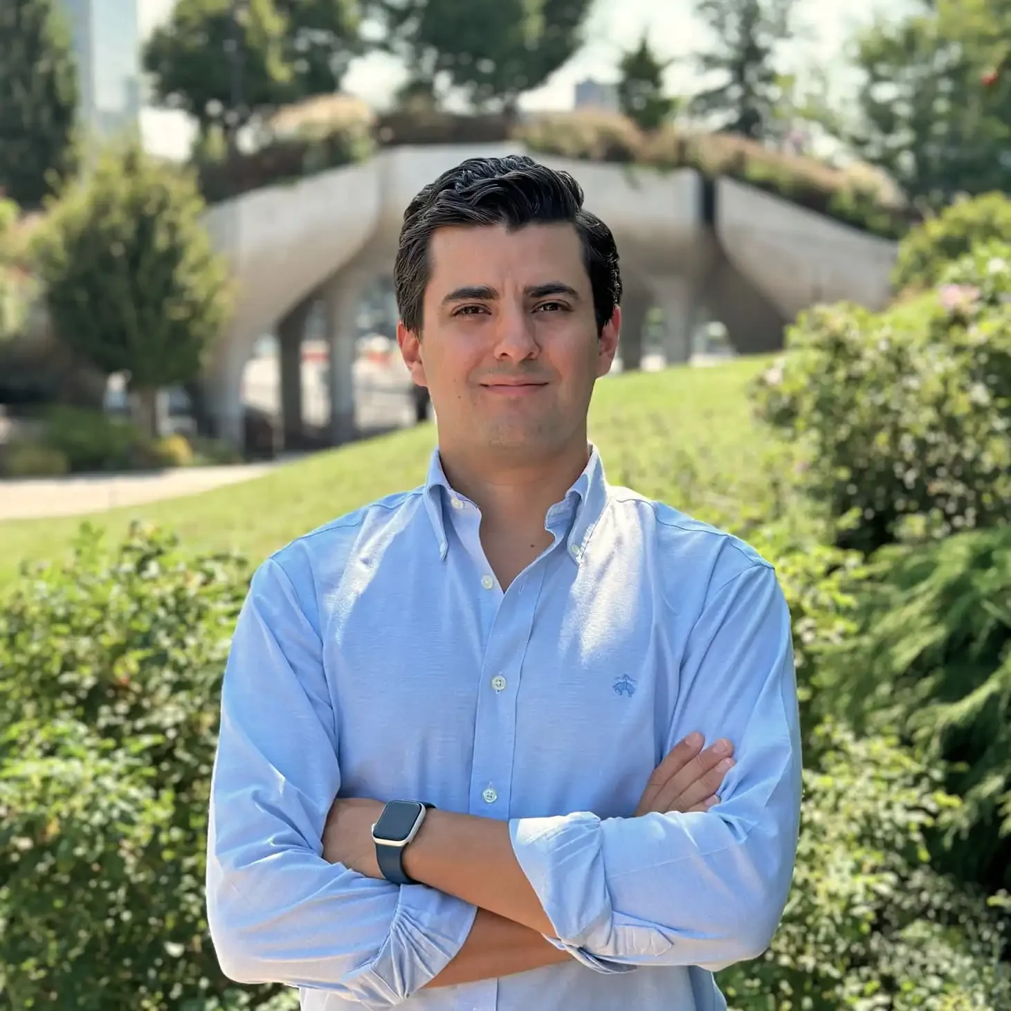 Un hombre joven con cabello oscuro y peinado hacia atrás, viste una camisa azul clara y lleva un reloj inteligente, de pie al aire libre en un parque con árboles y un puente en el fondo, con los brazos cruzados.